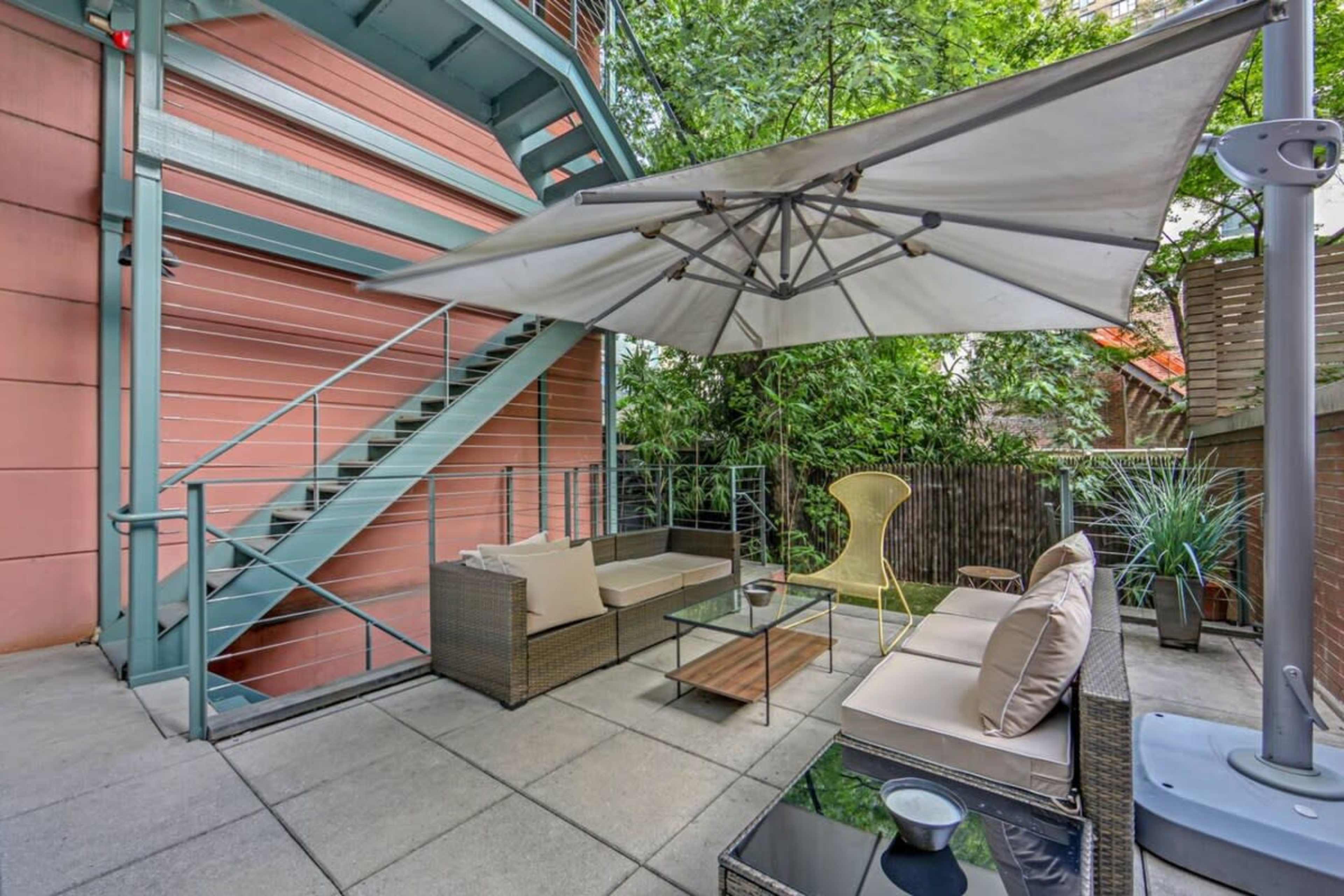 The image shows a patio area with a shaded seating arrangement, featuring a sectional couch, a coffee table, and potted plants, surrounded by greenery and a staircase in the background.