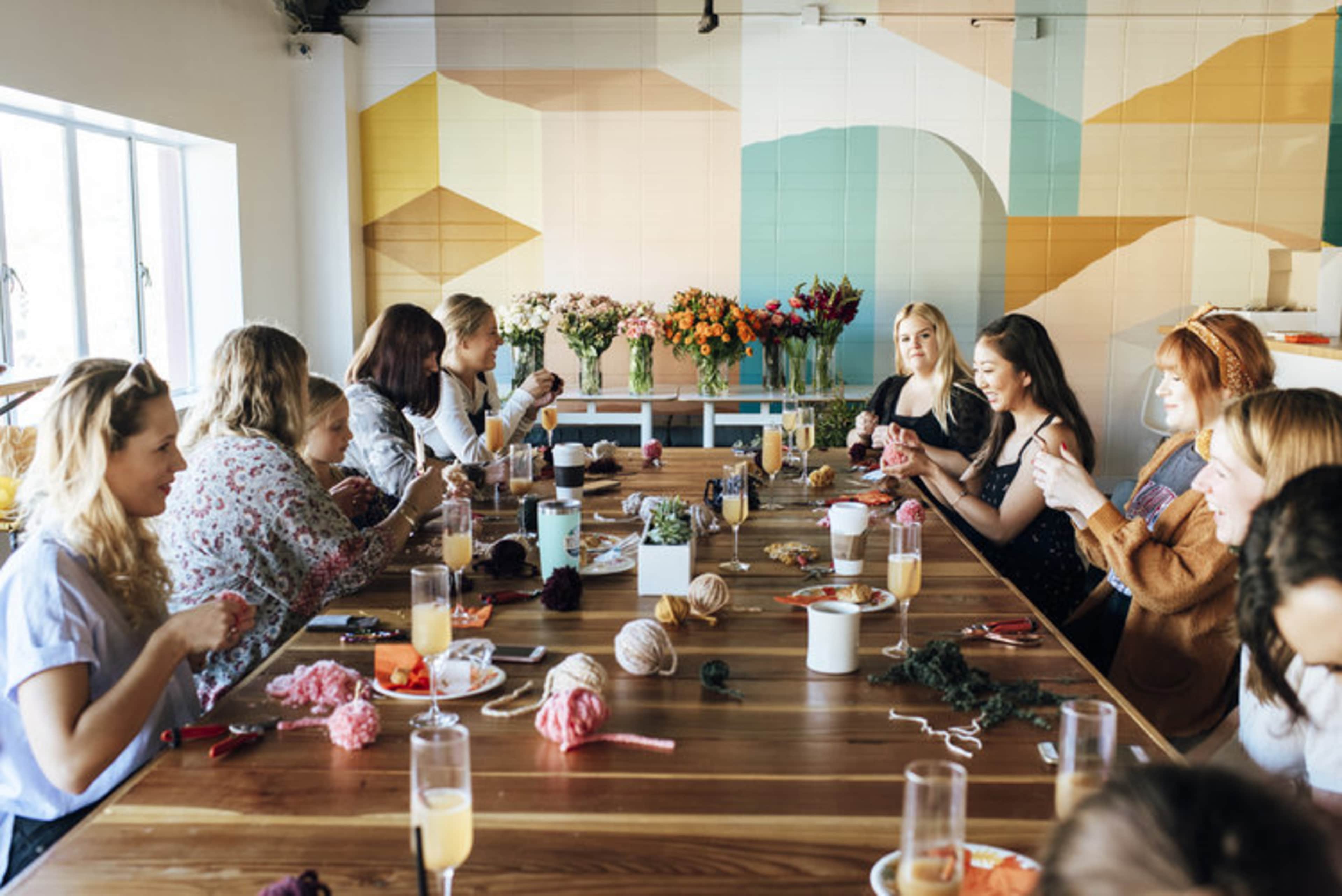 A group of people are seated around a long wooden table, engaged in a crafting activity with yarn and various materials, while colorful flowers are displayed in vases nearby.