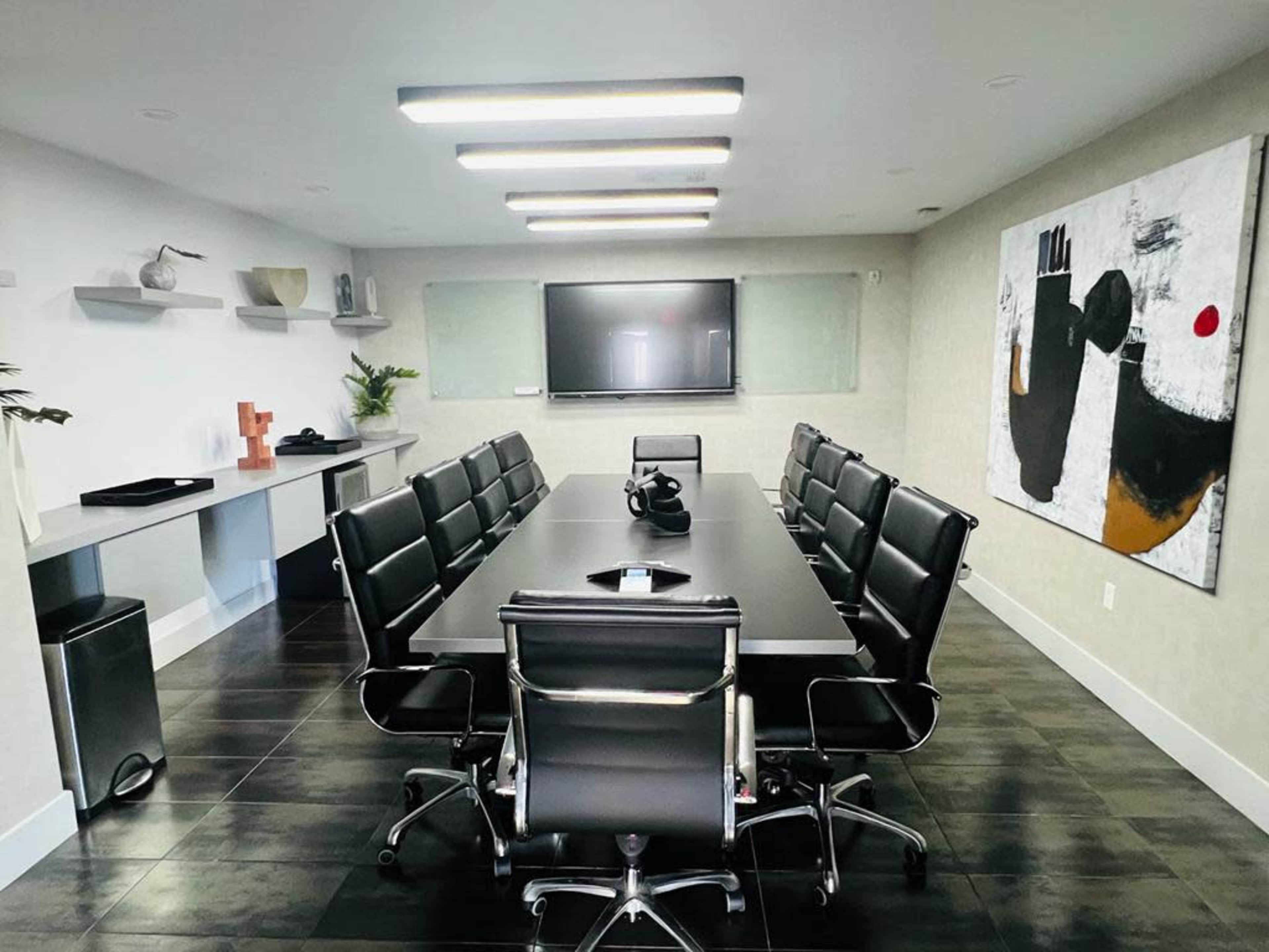 A conference room features a large rectangular table surrounded by black leather chairs, with a television mounted on the wall and a painting beside it.