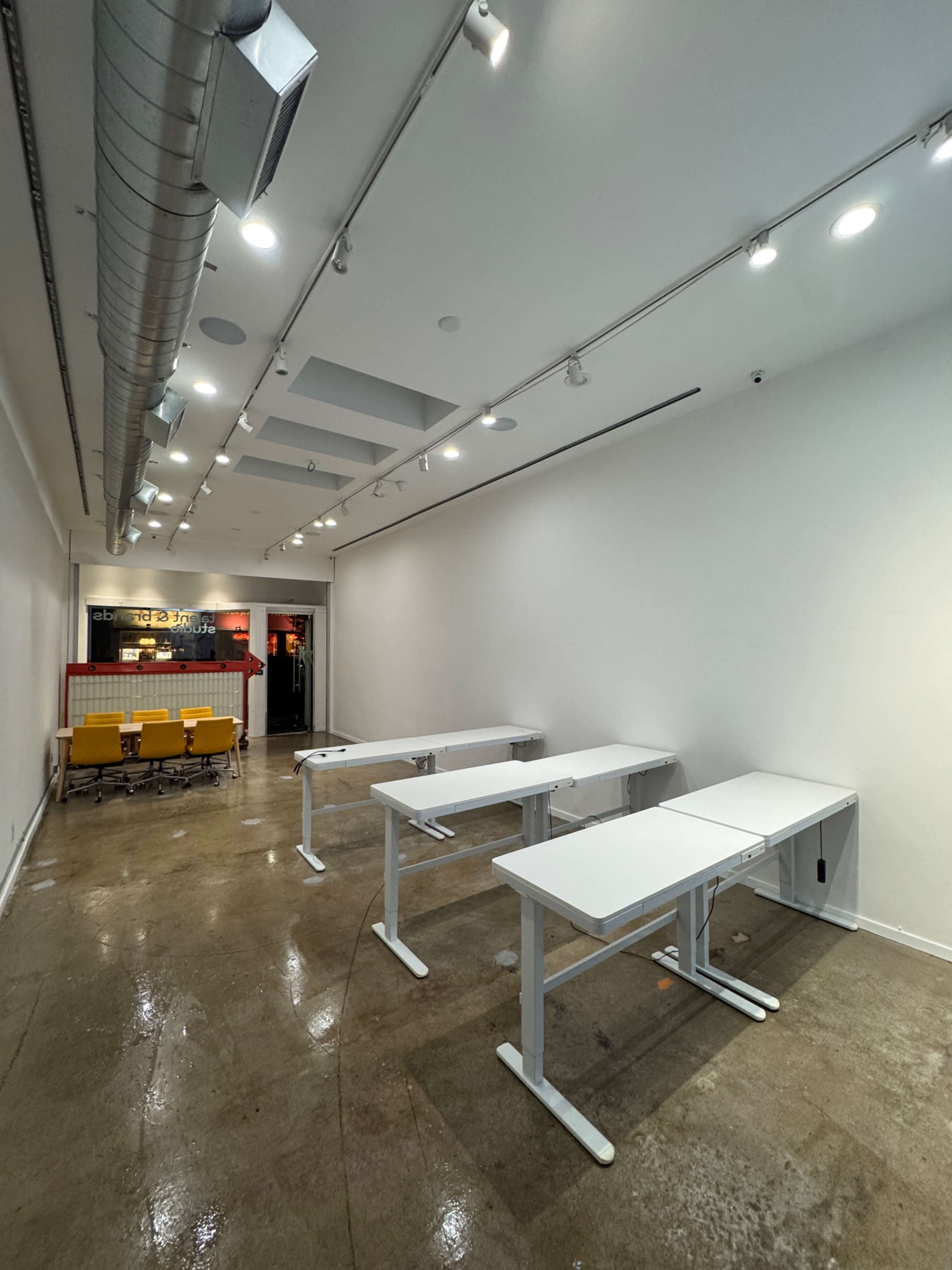 The image shows an empty room with white walls, polished concrete flooring, and several adjustable white desks arranged in a row.