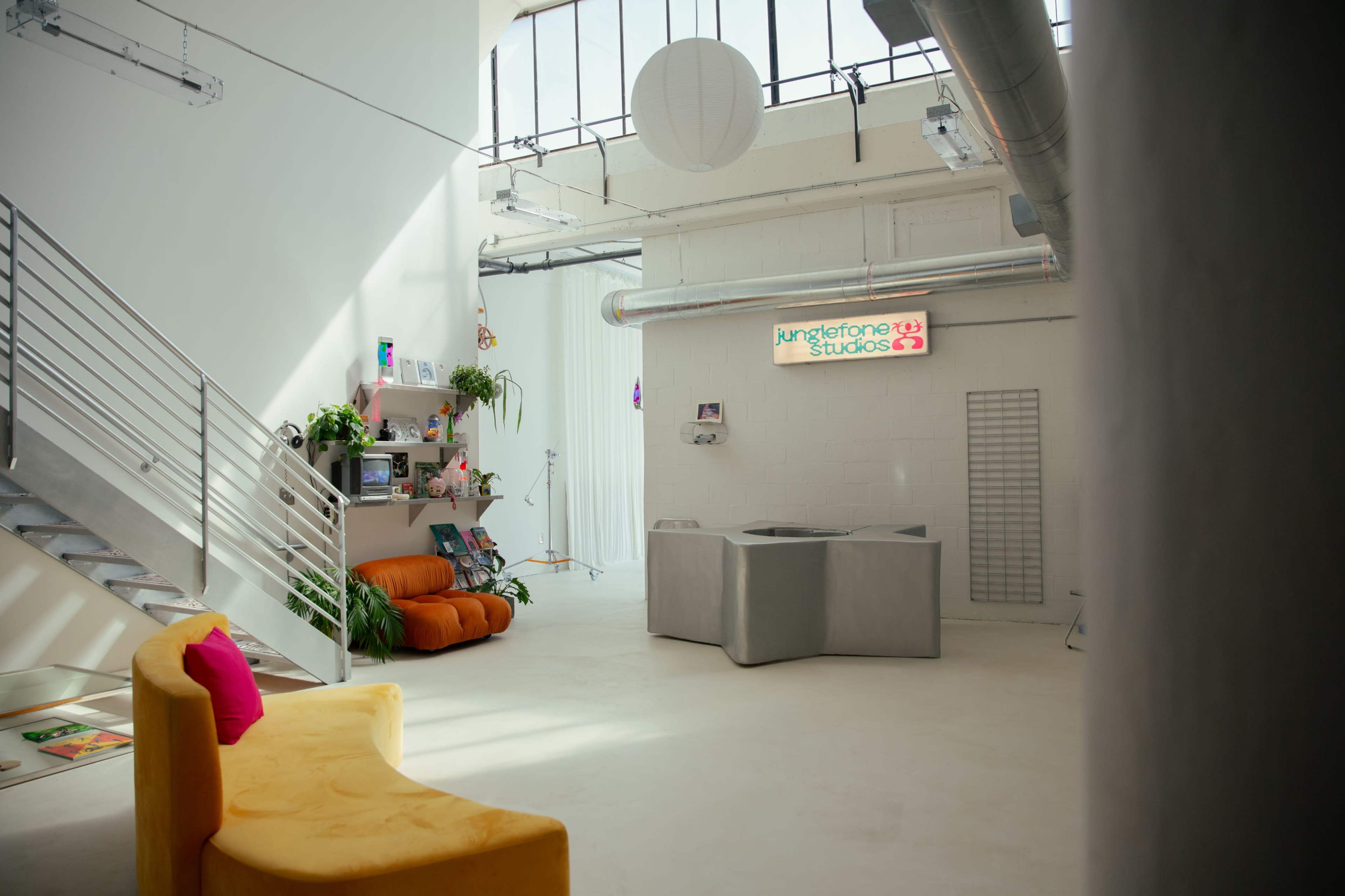 The image shows a modern studio interior featuring a staircase, colorful seating, and shelves with various items, illuminated by natural light from large windows.