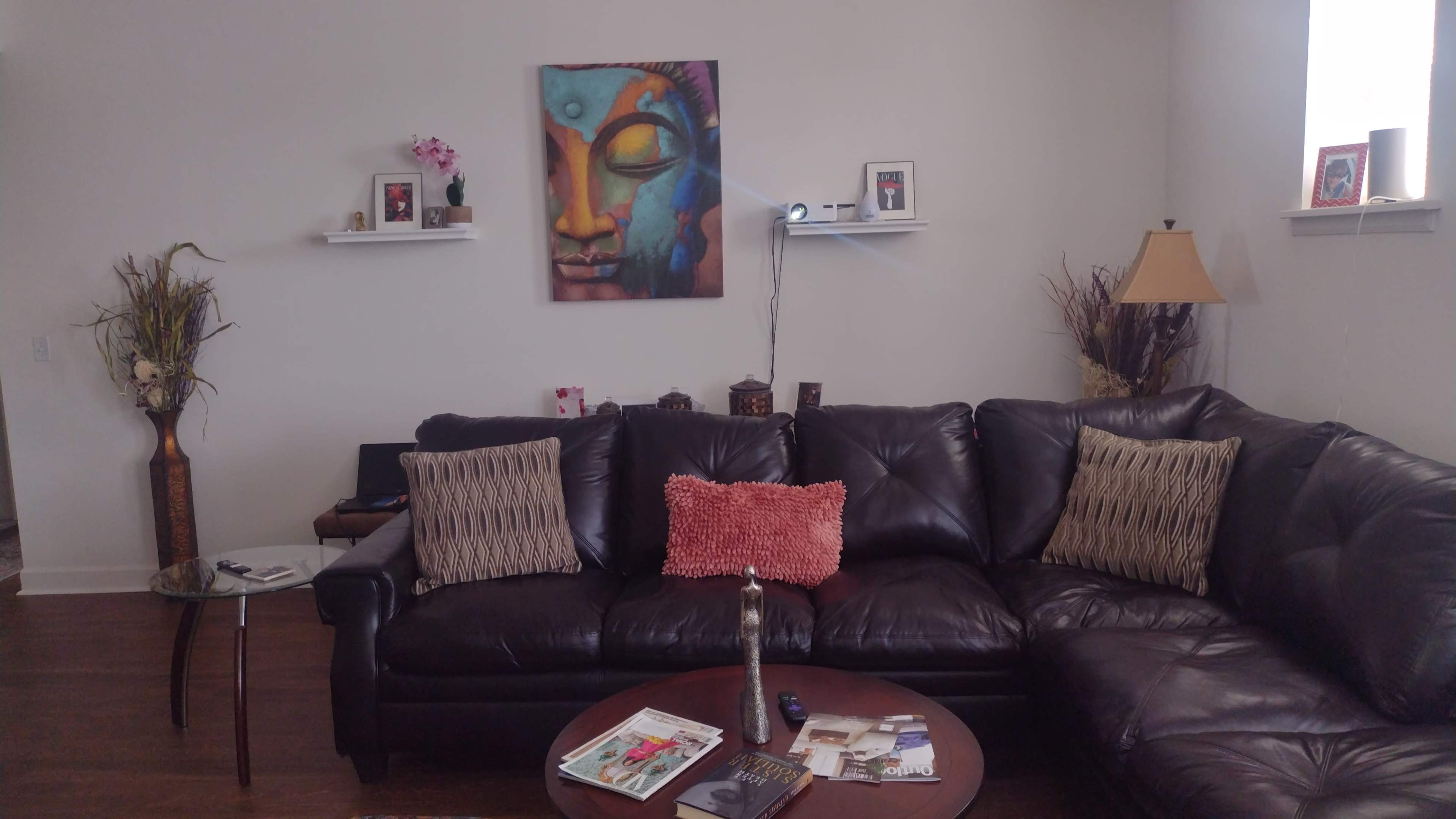The image shows a living room with a dark leather sectional sofa, a round coffee table with magazines, and a colorful Buddha painting on the wall above shelves holding decorative items.