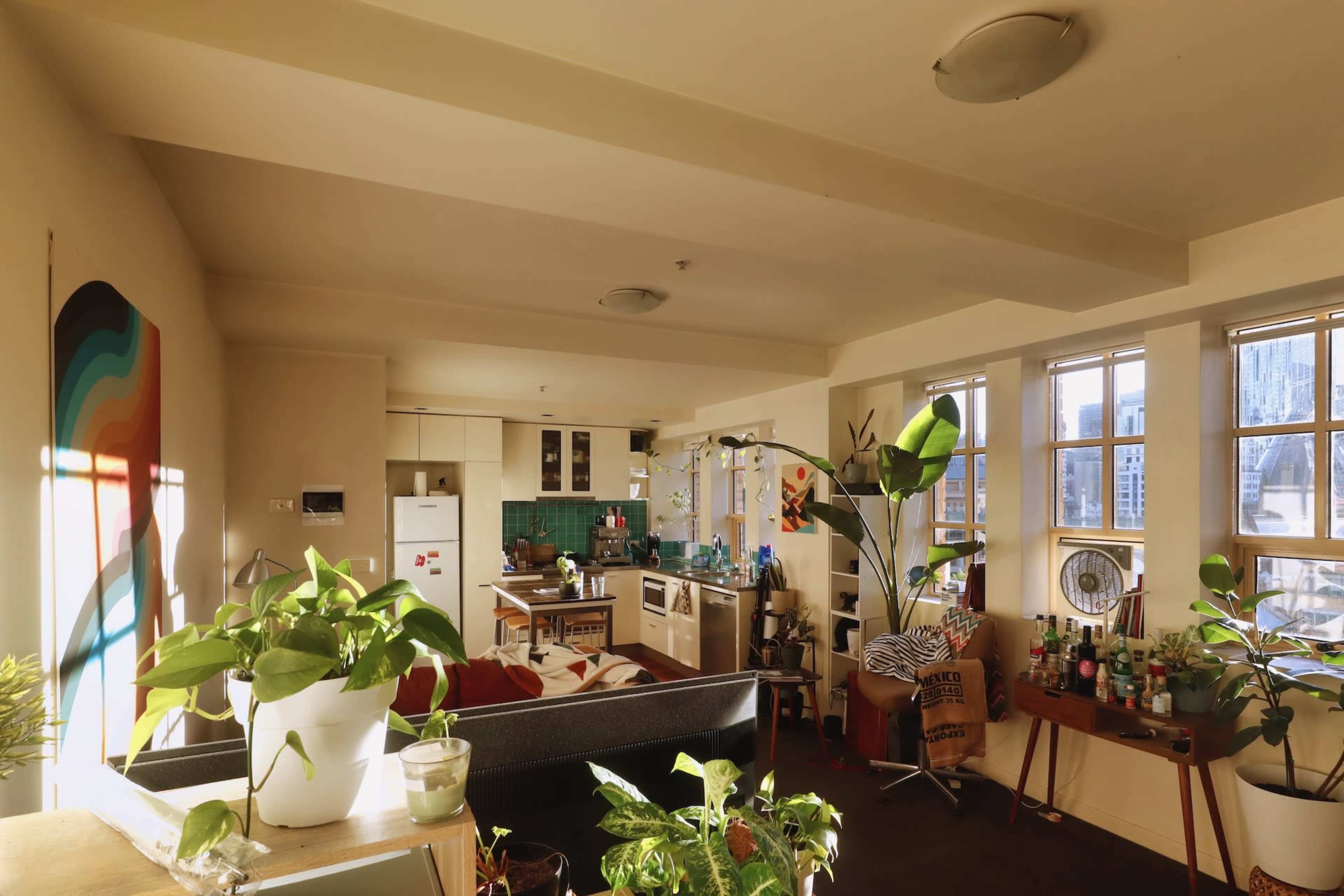 A bright, open-plan living space features a kitchen, a seating area with plants, and large windows showcasing a city view.