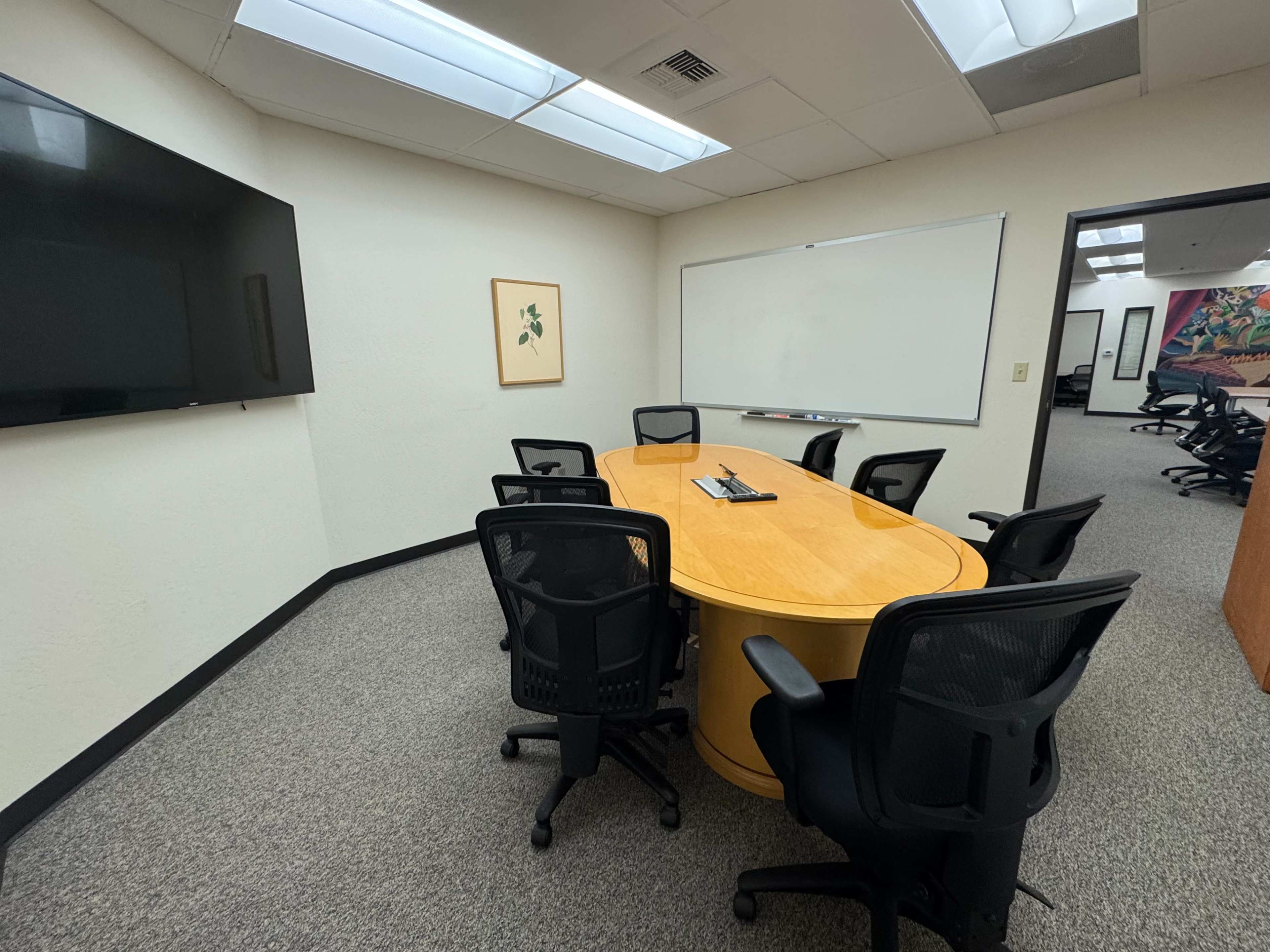 The image shows a conference room with a large oval table surrounded by six black ergonomic chairs, a whiteboard, and a large screen on one wall.