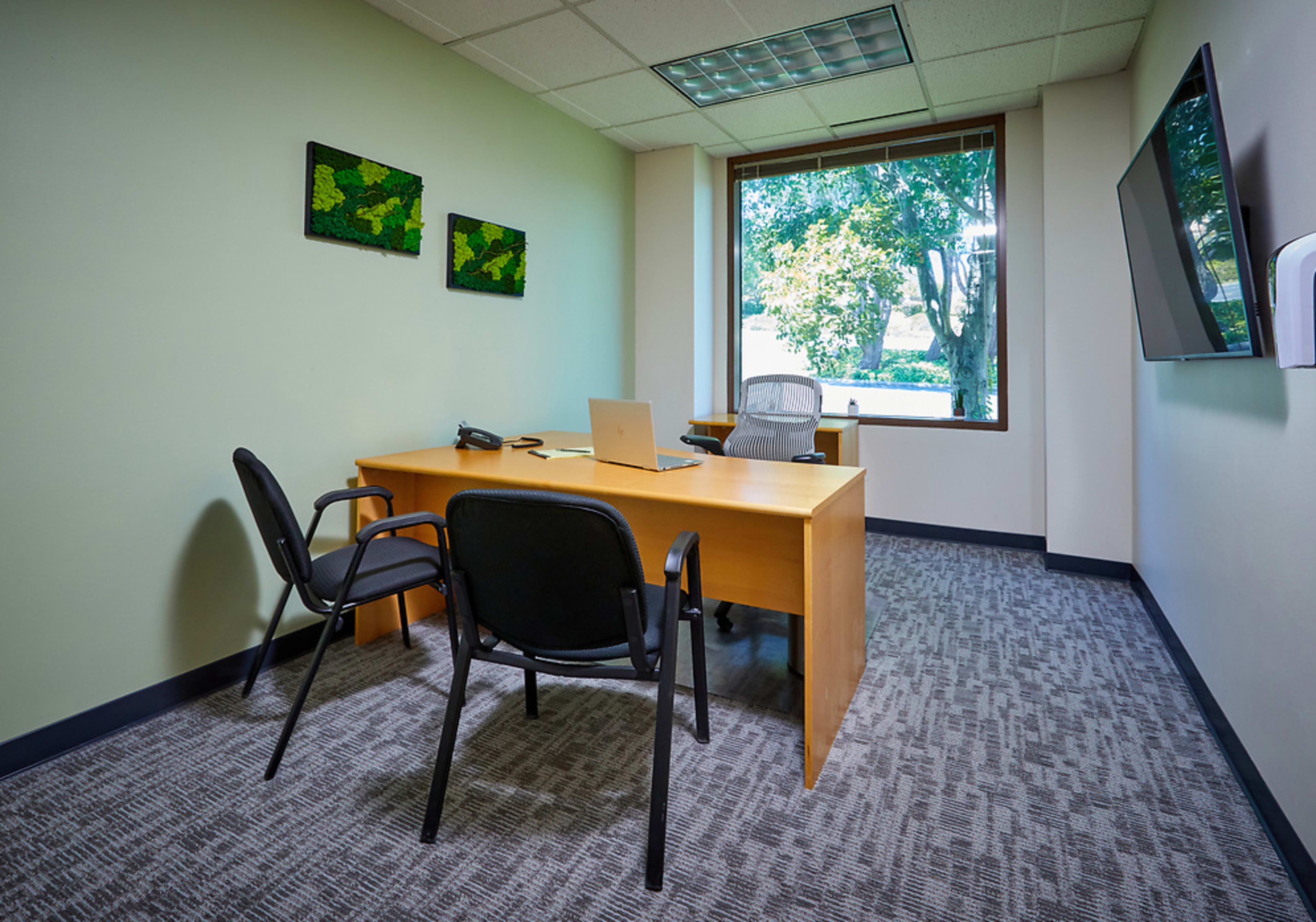 A small office with a wooden desk, two black chairs, a computer, and a television mounted on the wall, all with a window view of greenery outside.