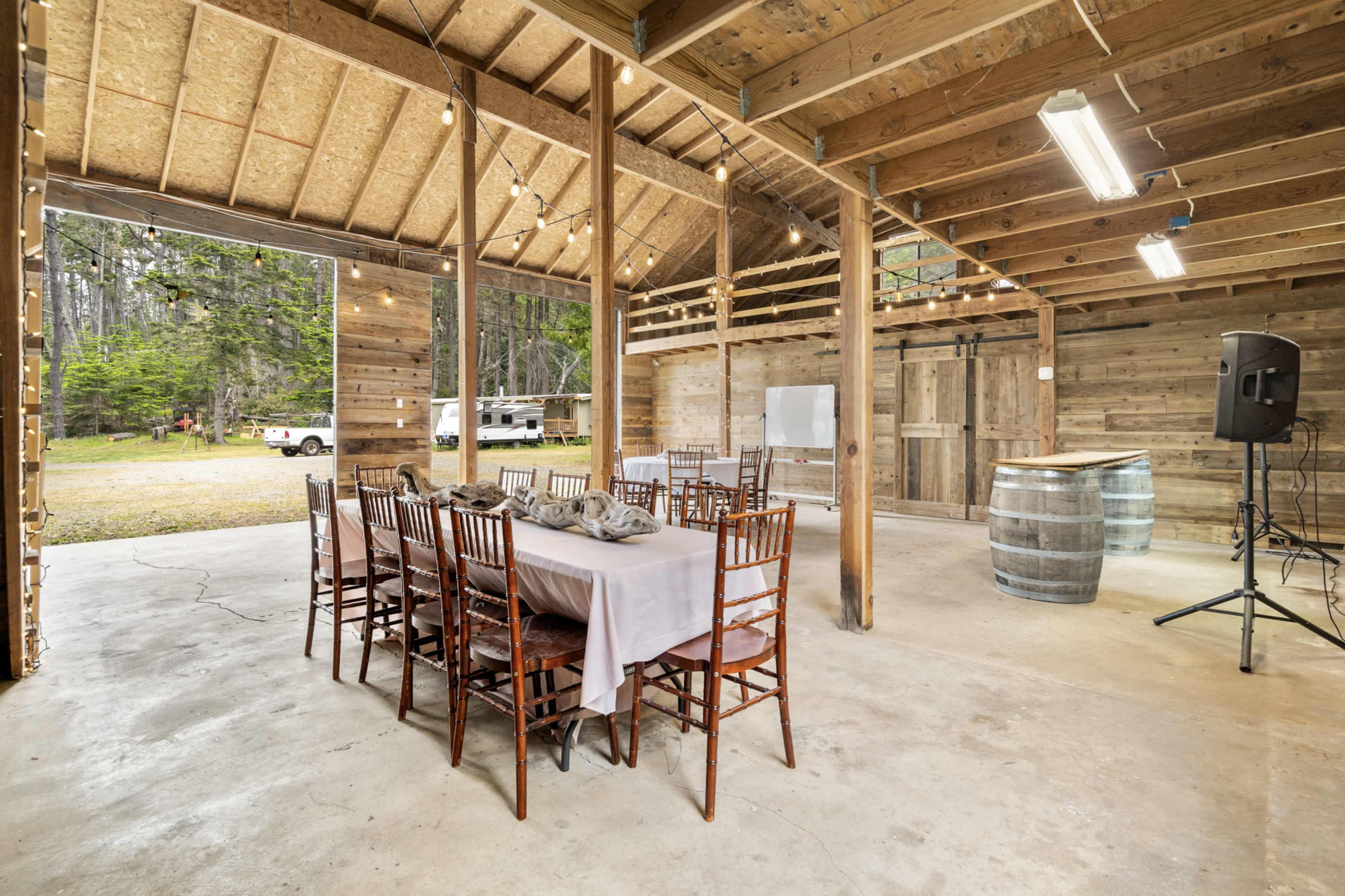 The image shows a rustic wooden interior of a barn-like space with a long dining table set for a gathering, surrounded by wooden chairs and string lights overhead.