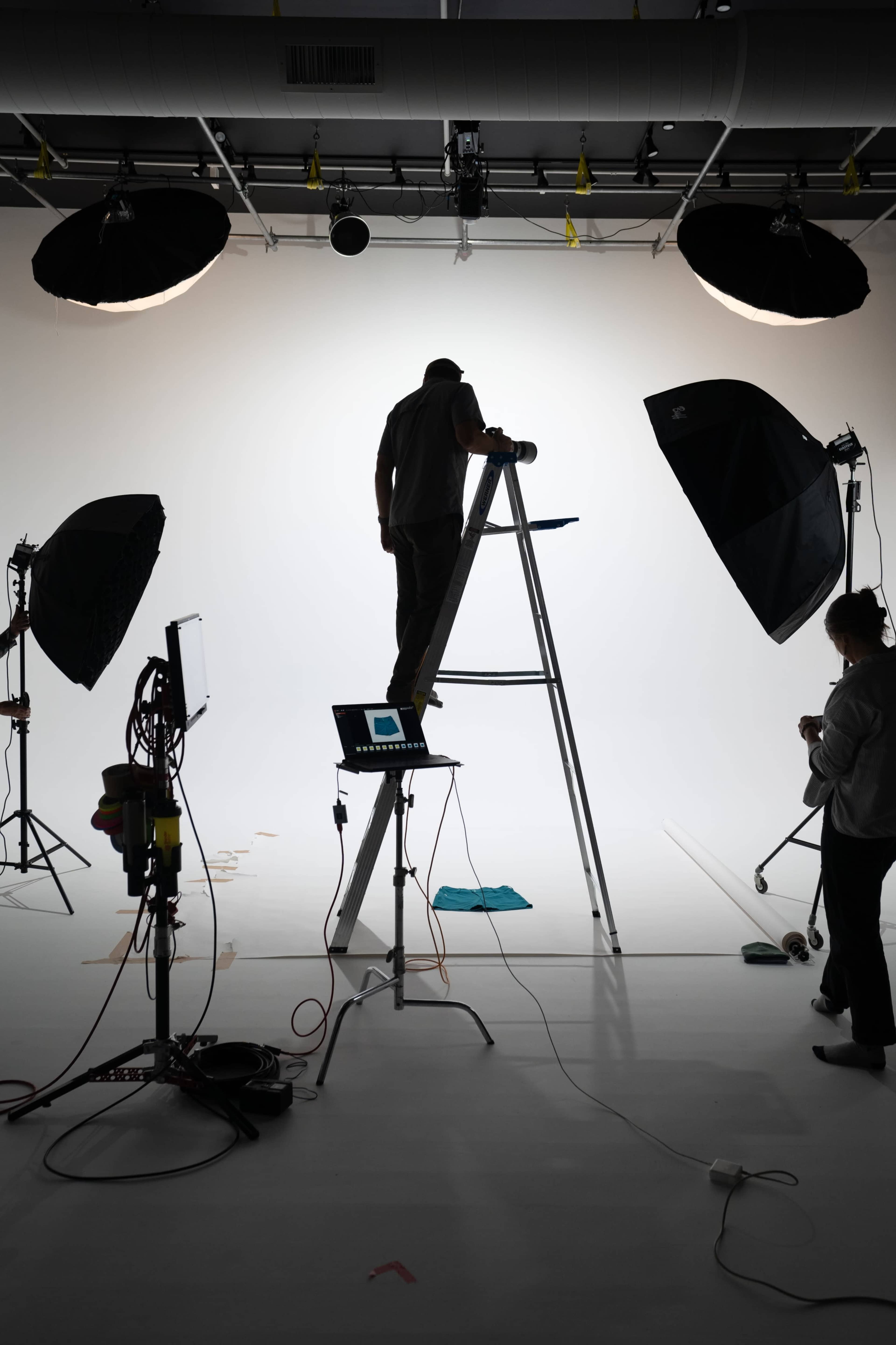 A person stands on a ladder within a studio, adjusting lighting equipment while another individual prepares materials nearby.