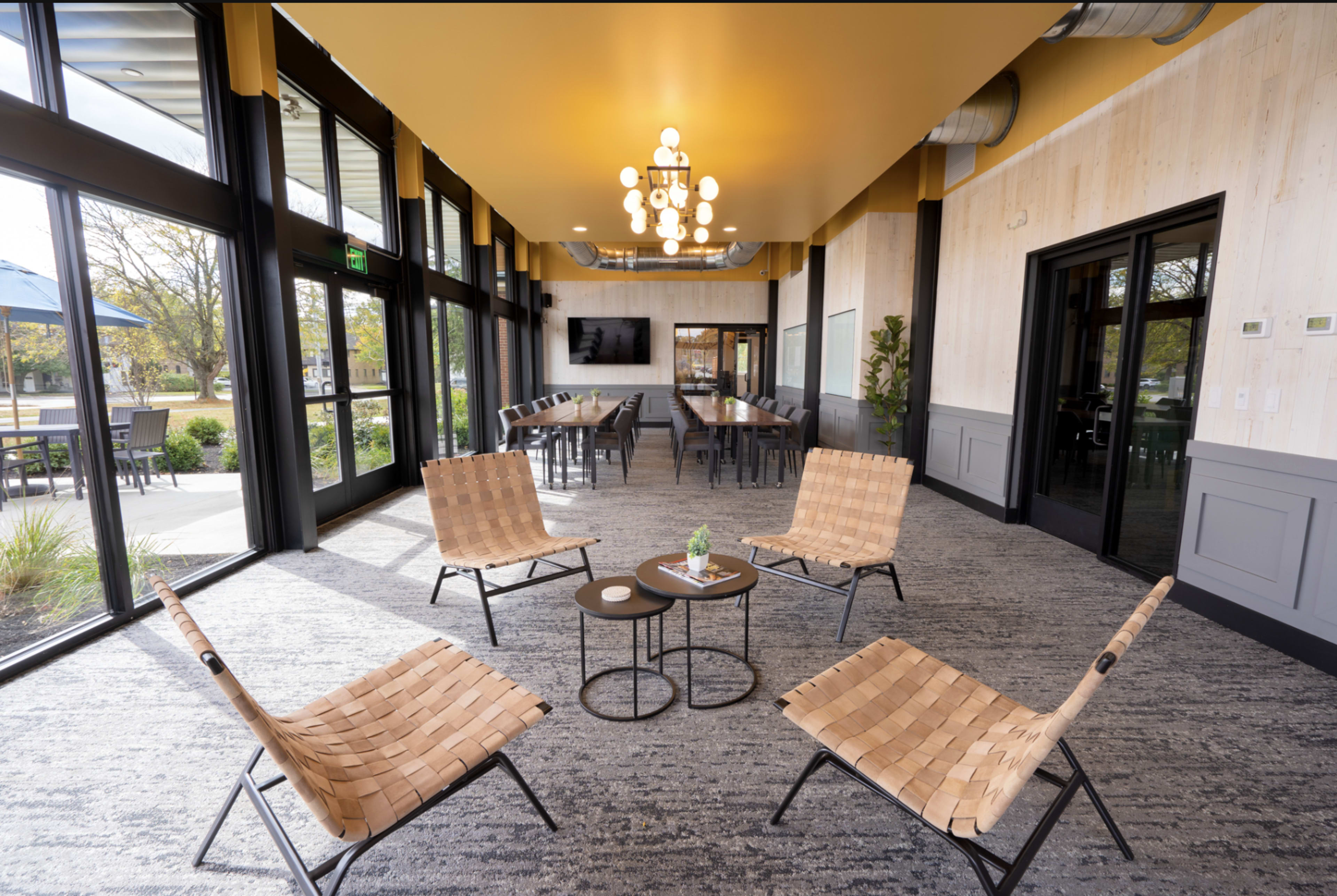 The image shows a modern indoor meeting space featuring four woven chairs arranged around a small table, illuminated by a chandelier and surrounded by large windows.