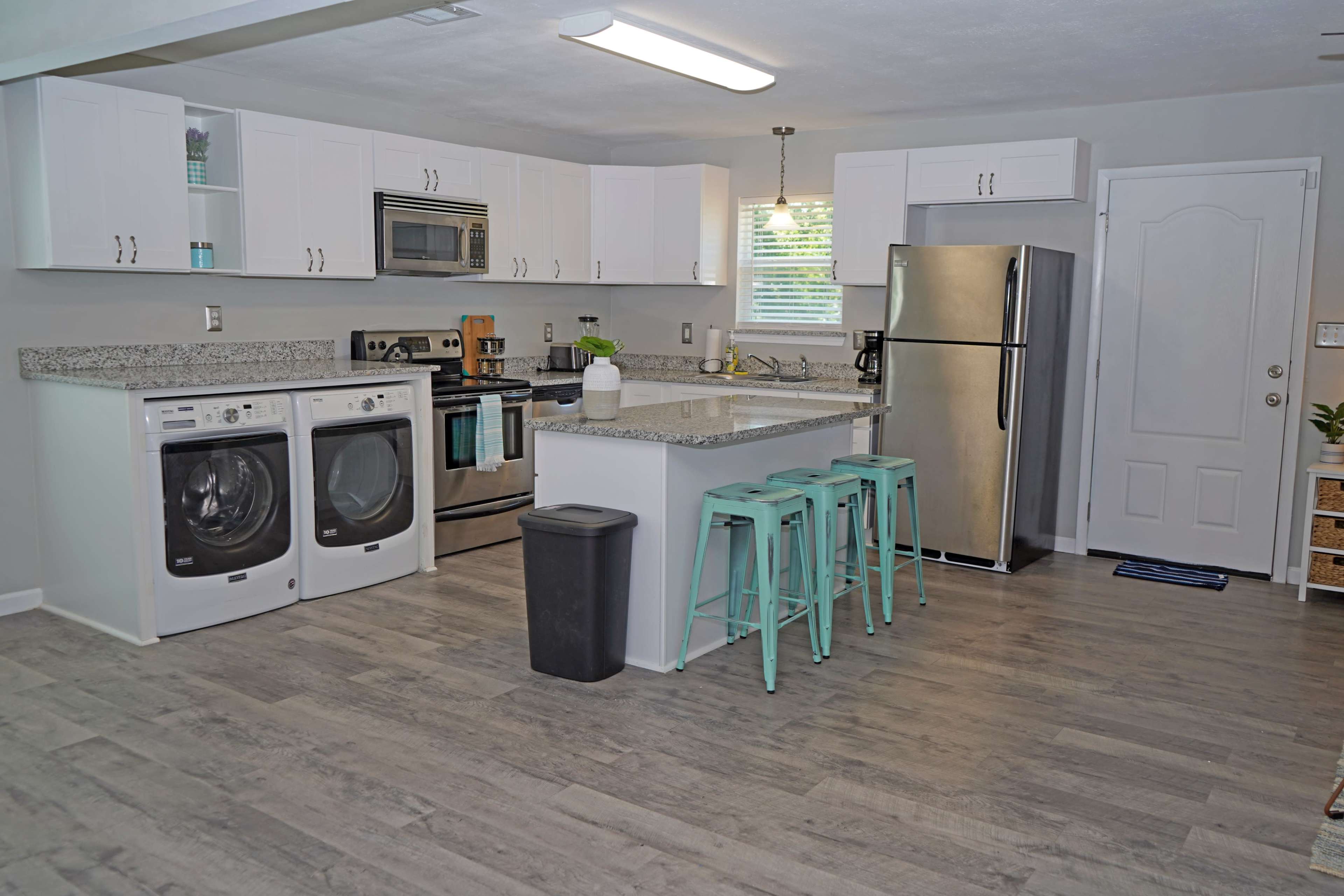 A modern kitchen features a washer and dryer, stainless steel appliances, a centrally located island with turquoise bar stools, and light-colored cabinetry.