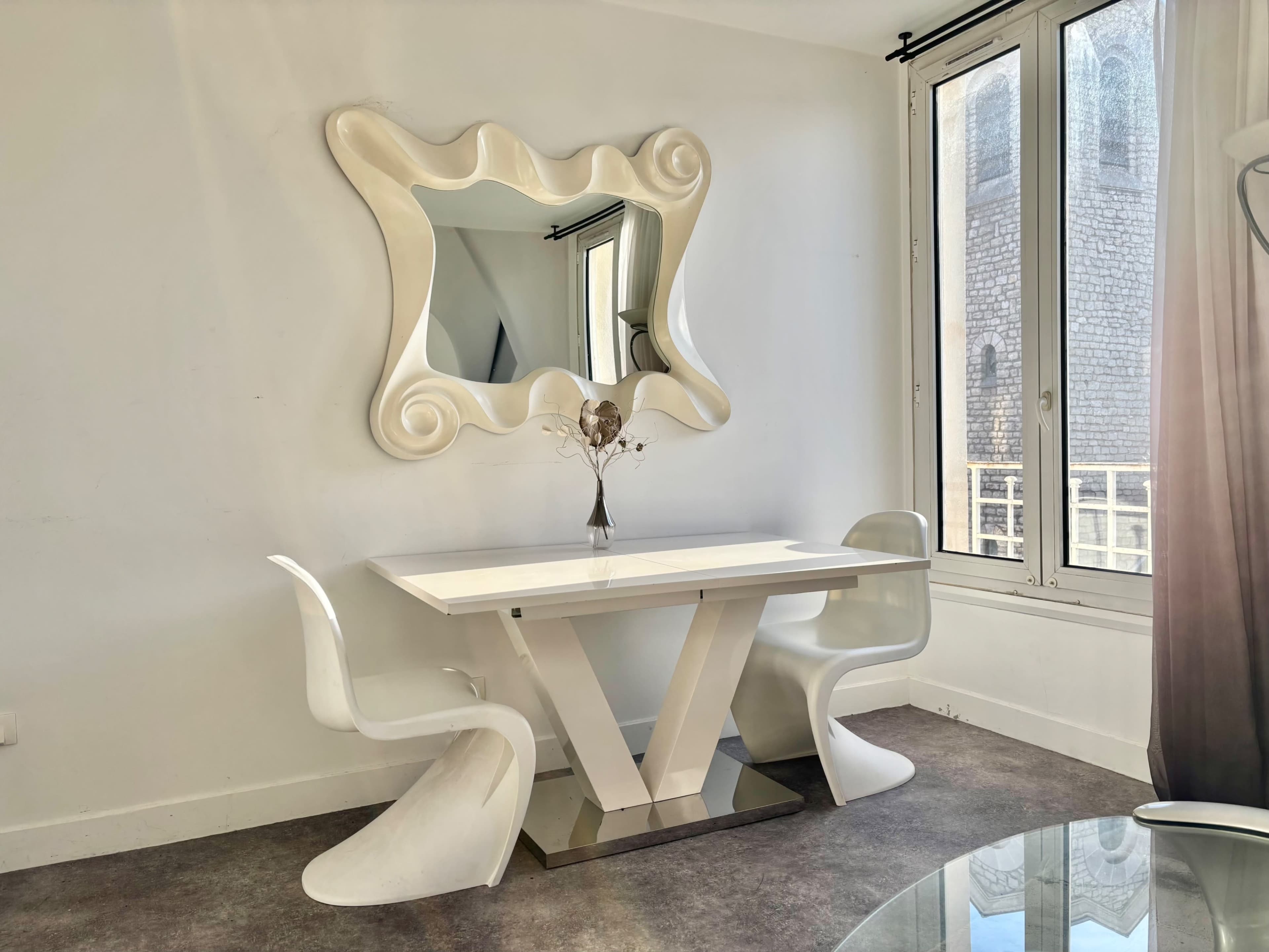 A modern dining table with two white chairs is positioned near large windows, reflecting a decorative, wavy mirror on the wall.