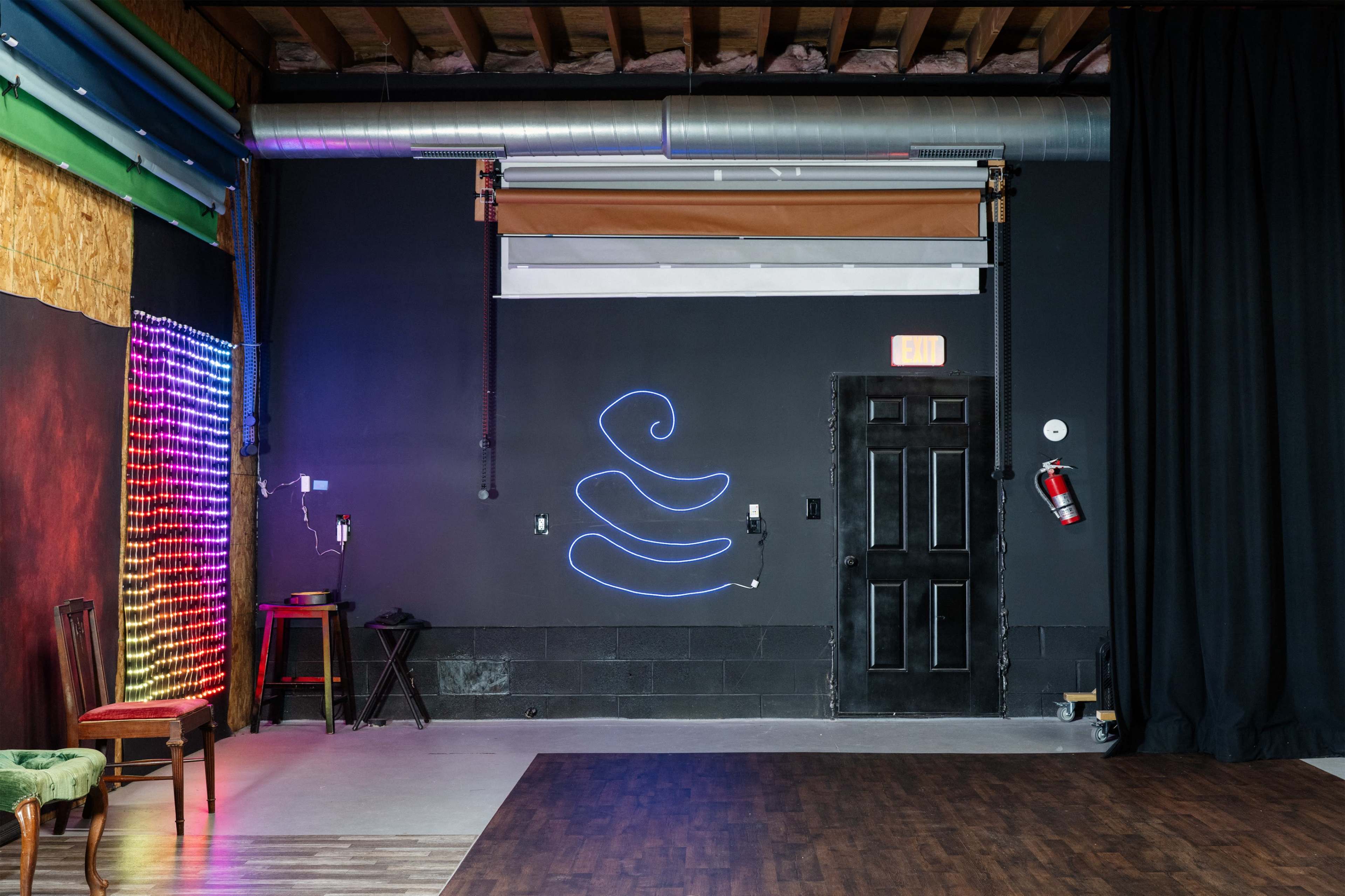 The image shows an indoor space featuring a dark wall with a neon design, a fire extinguisher near a black door, and an illuminated wall panel emitting colorful light.
