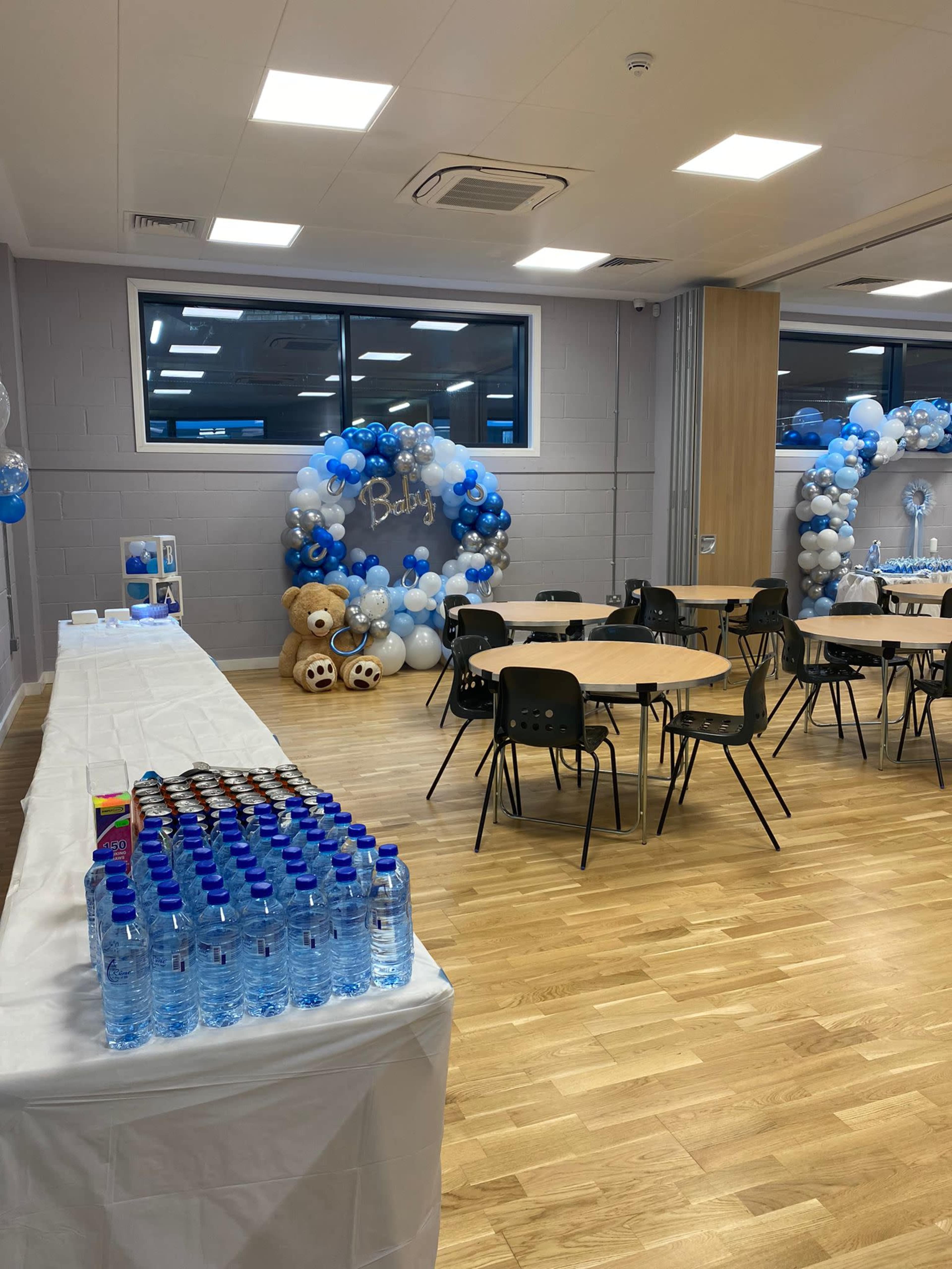 The room is set up for a baby shower, featuring tables with chairs, decorative balloons, bears, and a long table with bottled water.