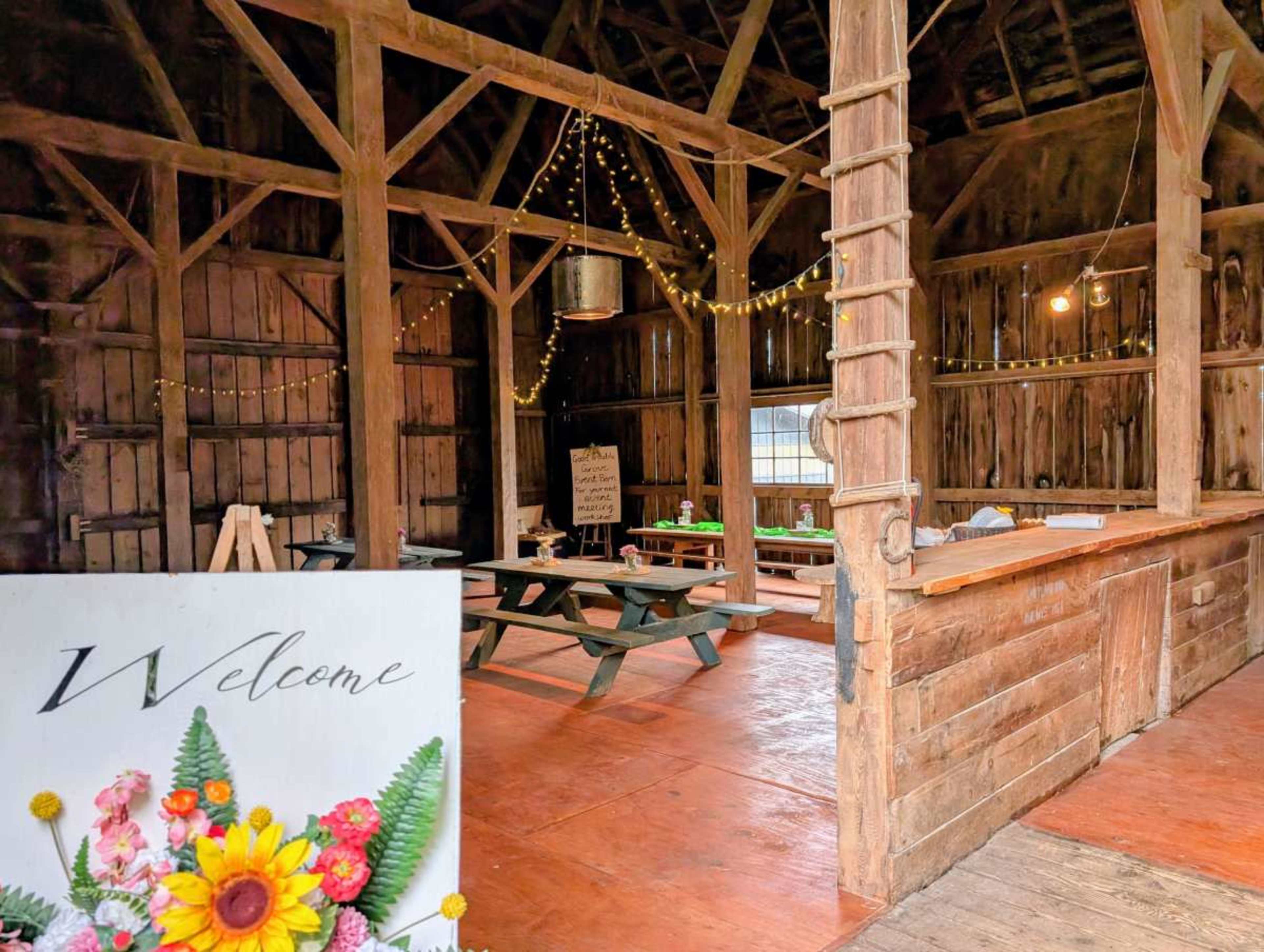 The interior of a rustic barn features wooden beams, tables, and decorative lighting, with a "Welcome" sign adorned with flowers prominently featured in the foreground.