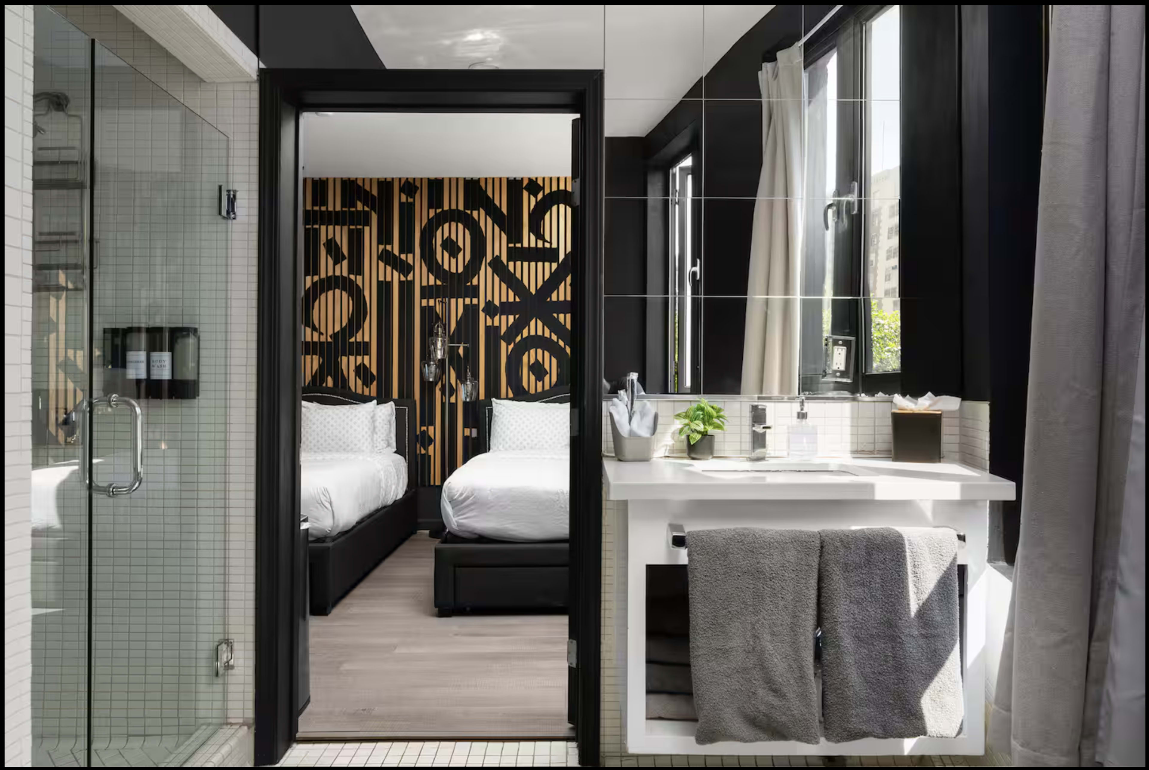 A modern bathroom features a glass shower and a view into a bedroom with patterned wallpaper and two beds.