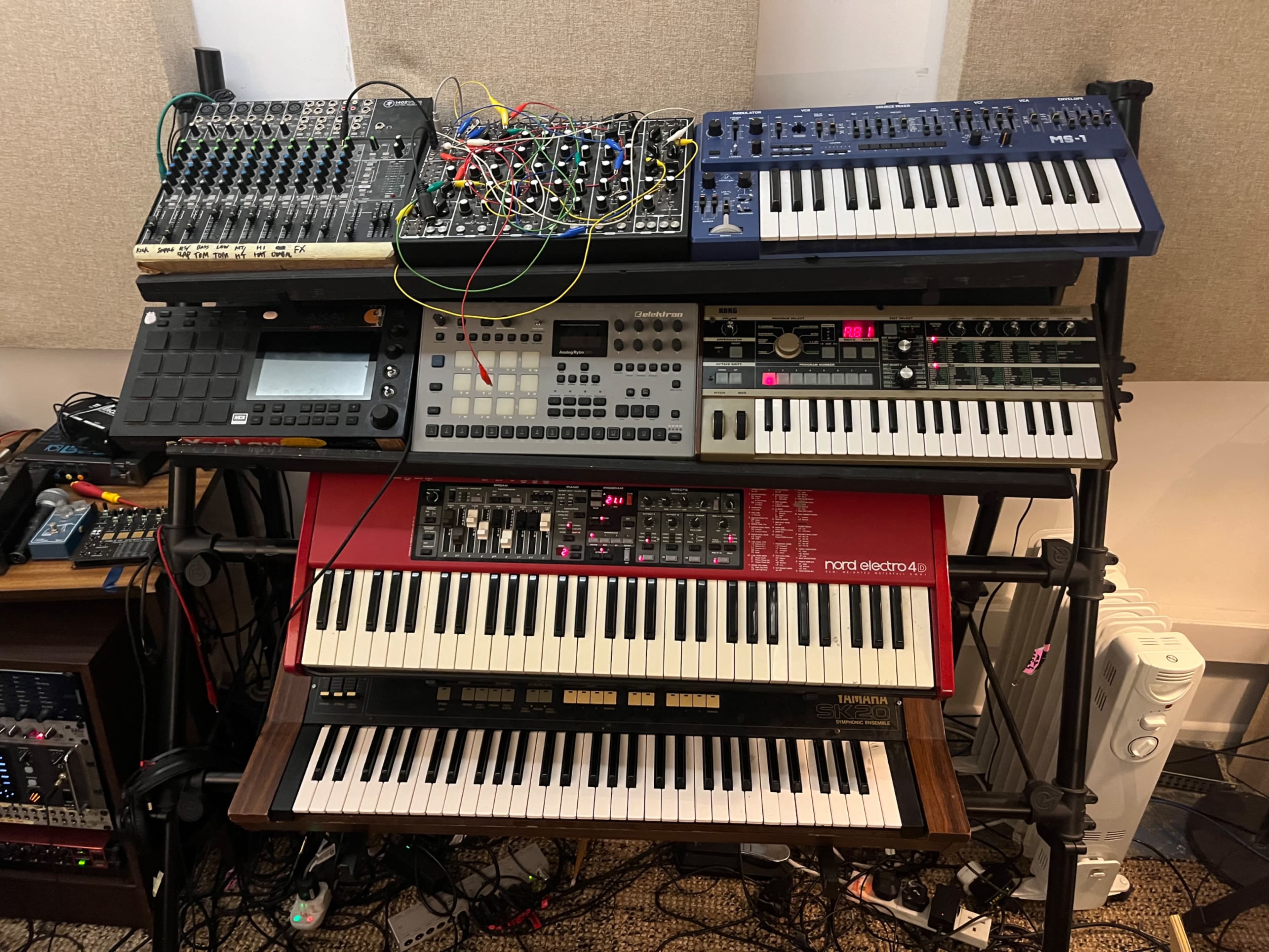 The image shows a music production setup featuring multiple synthesizers, a mixing console, and various electronic instruments arranged on a multi-tiered stand.