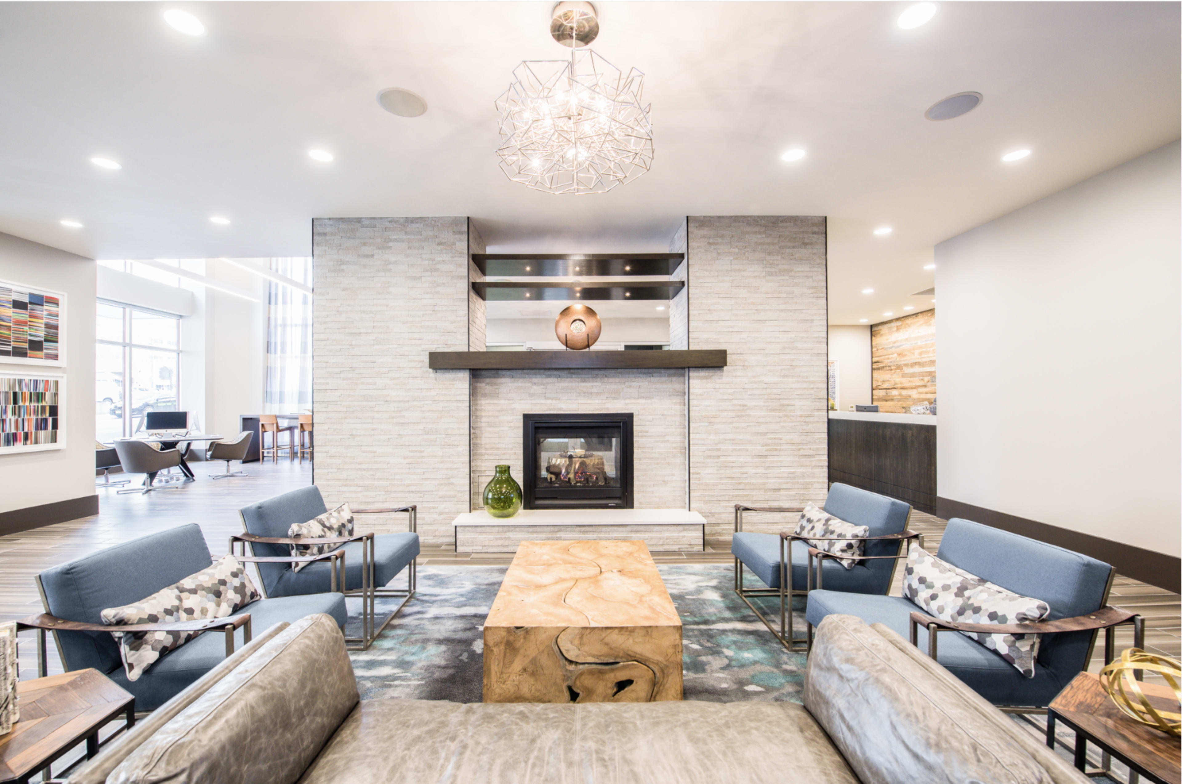 The image features a modern lobby with blue upholstered chairs, a wooden coffee table, a stone fireplace with a decorative piece, and a well-lit, open design.