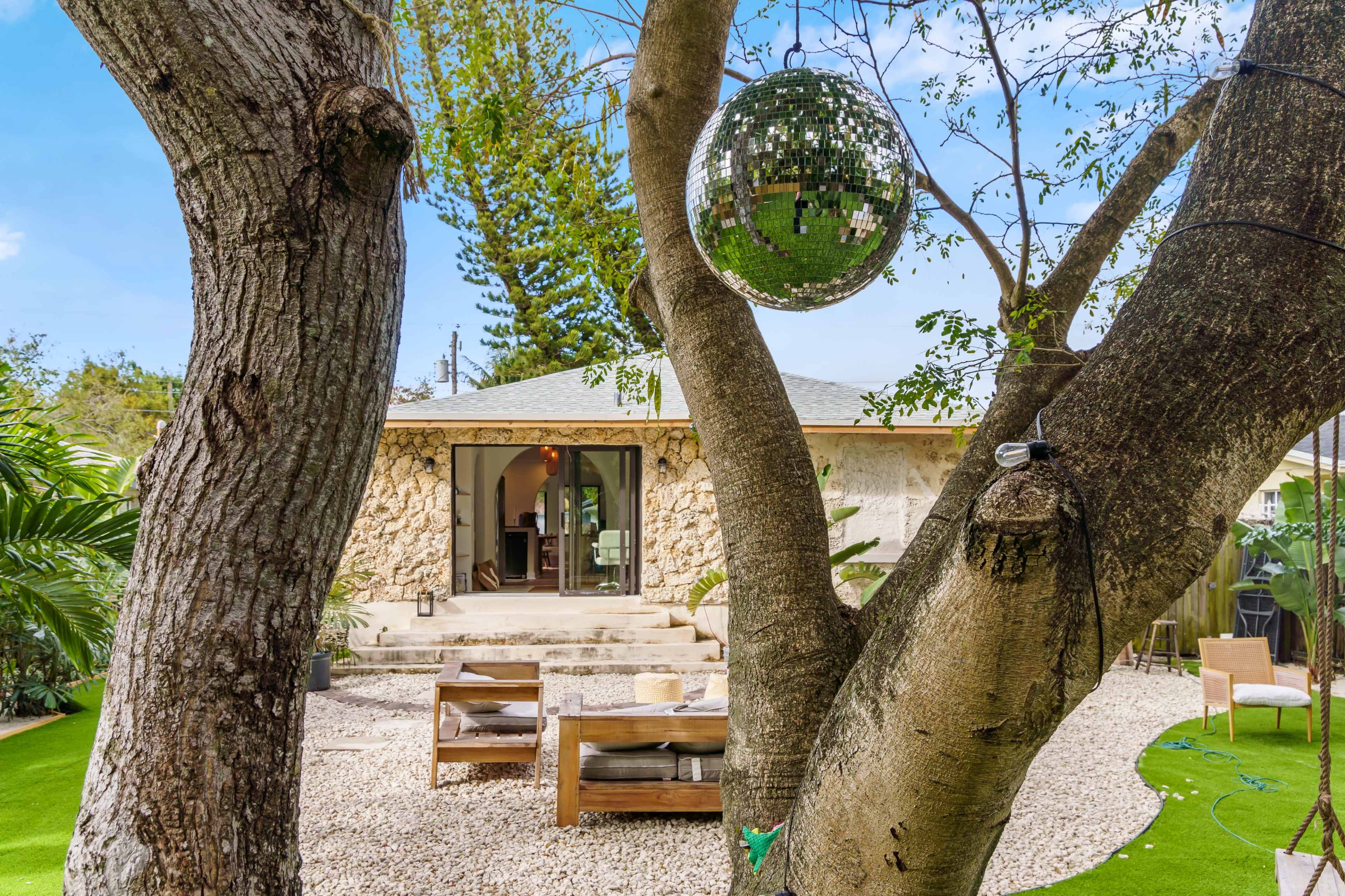 A backyard features a stone house with a patio, surrounded by greenery and highlighted by a disco ball hanging from a tree.