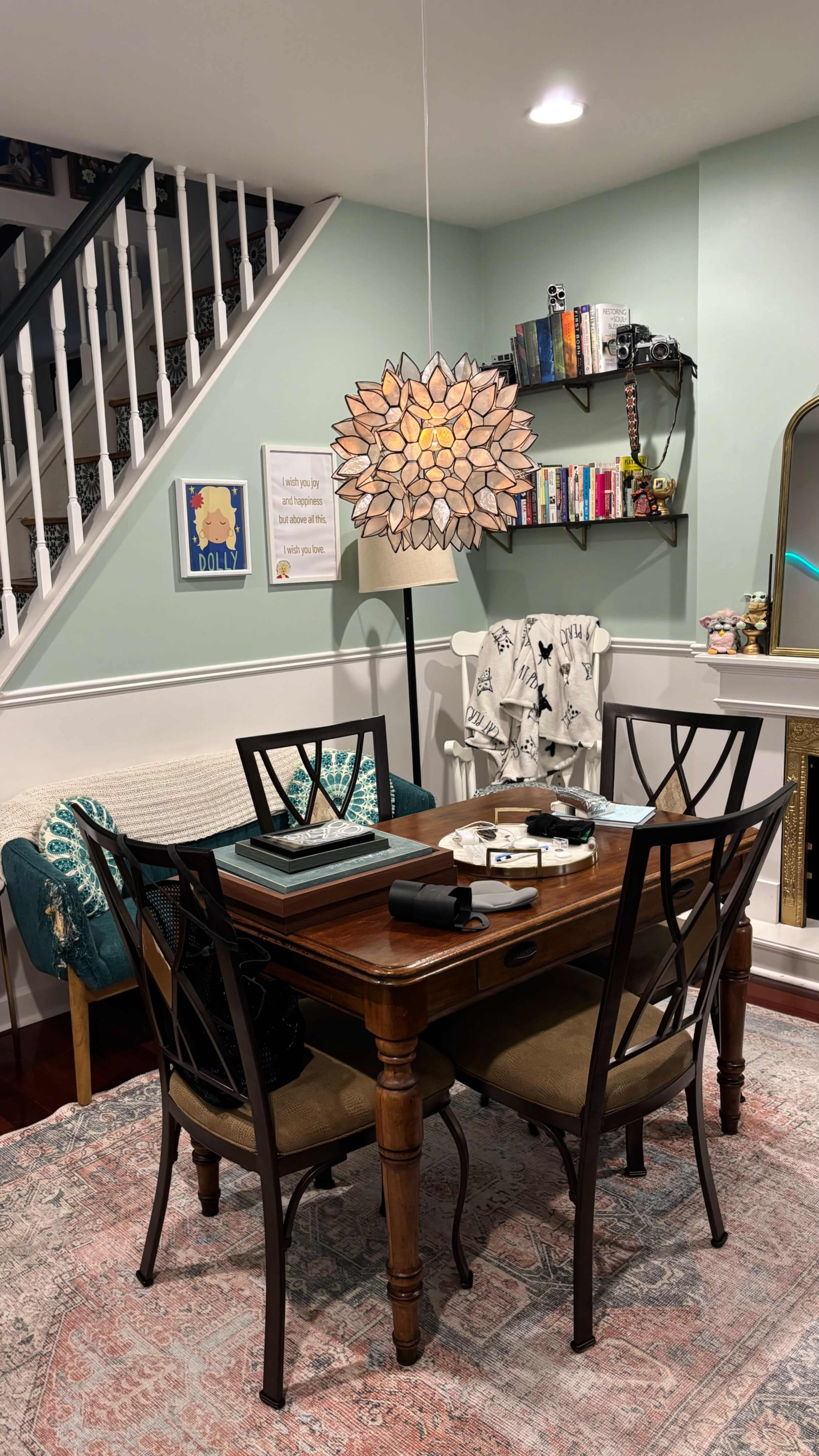 A dining area features a wooden table with four chairs, a decorative light fixture above, and a bookshelf along the wall.