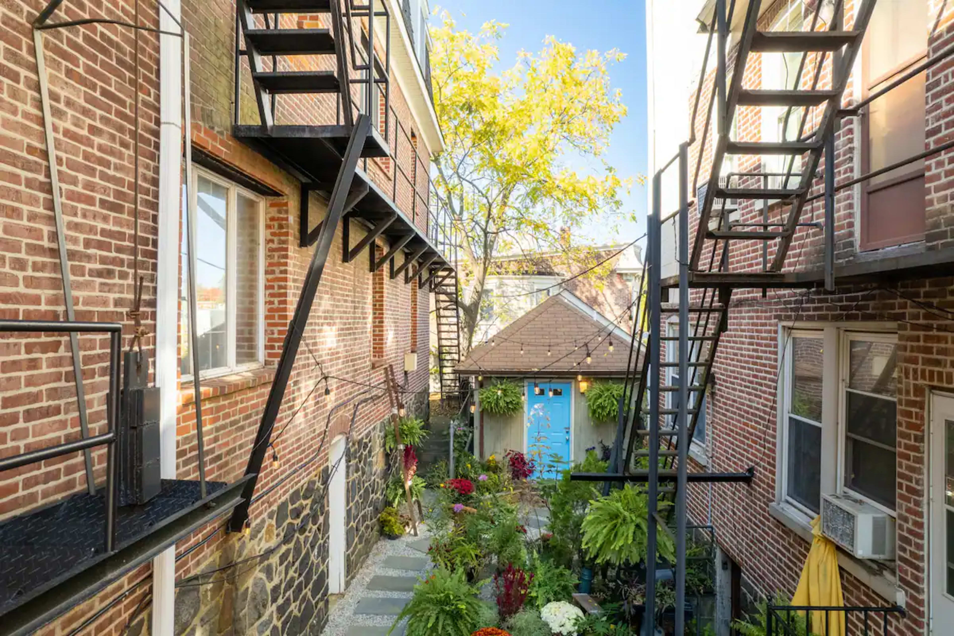 A narrow alley between two brick buildings, featuring metal fire escapes and a small garden with colorful plants leading to a blue door.