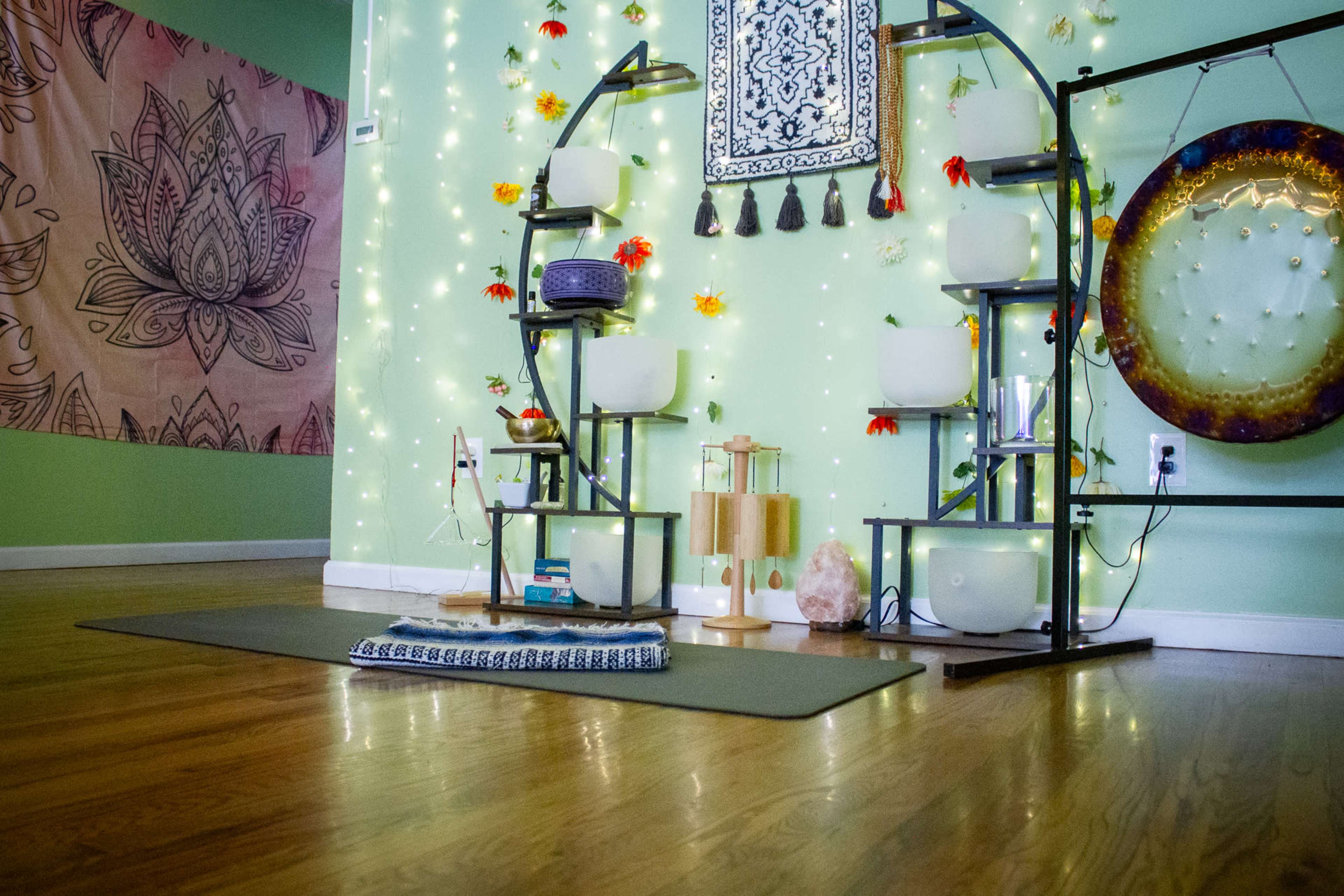 The image shows a tranquil room with a yoga mat on the floor, surrounded by decorated shelves with sound bowls and plants, illuminated by soft string lights.