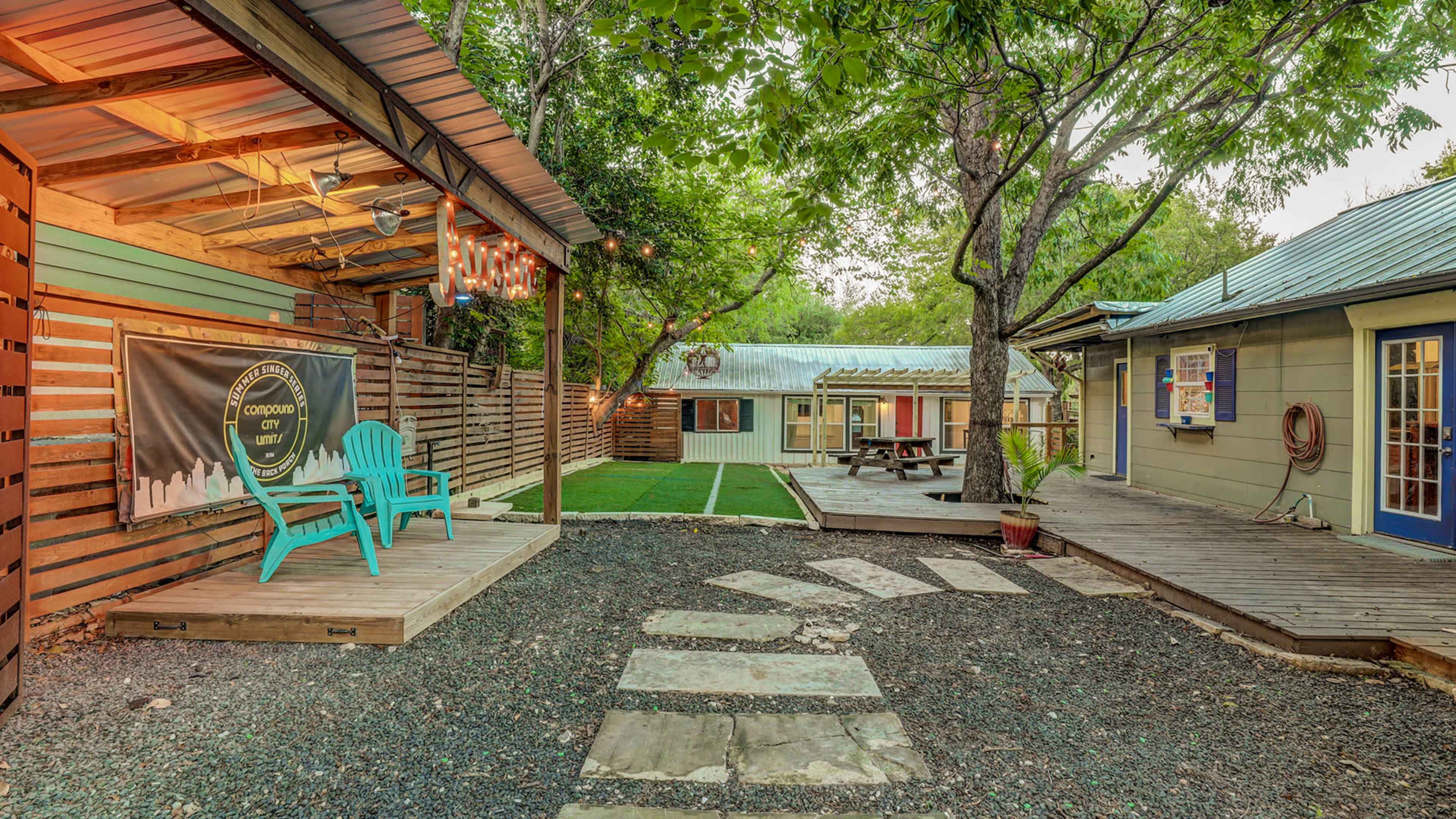 A landscaped outdoor space featuring a stone pathway, a grassy area with picnic tables, and two separate buildings surrounded by trees.