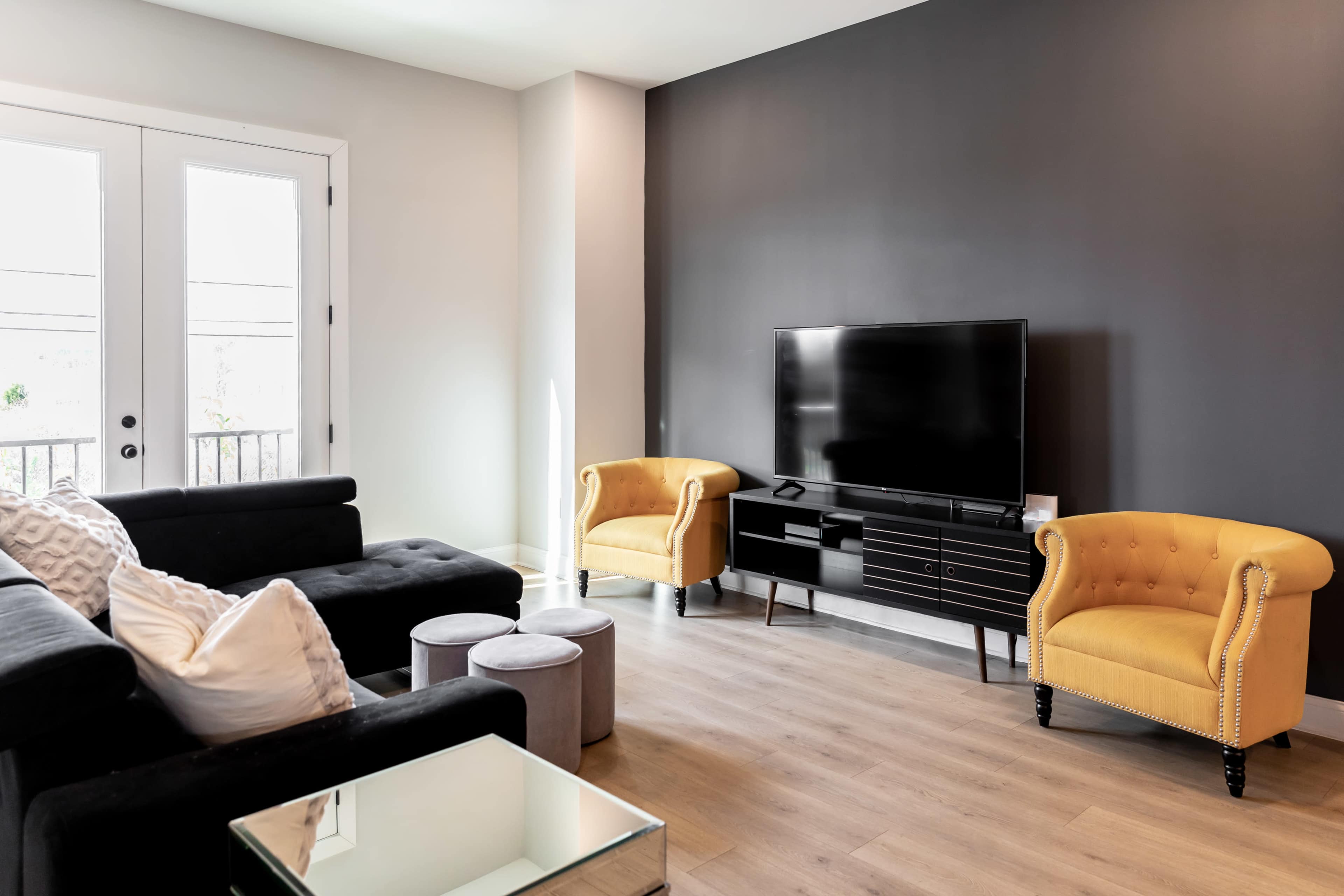 A modern living room features a black sofa, yellow accent chairs, a black television stand with a TV, and light wood flooring.