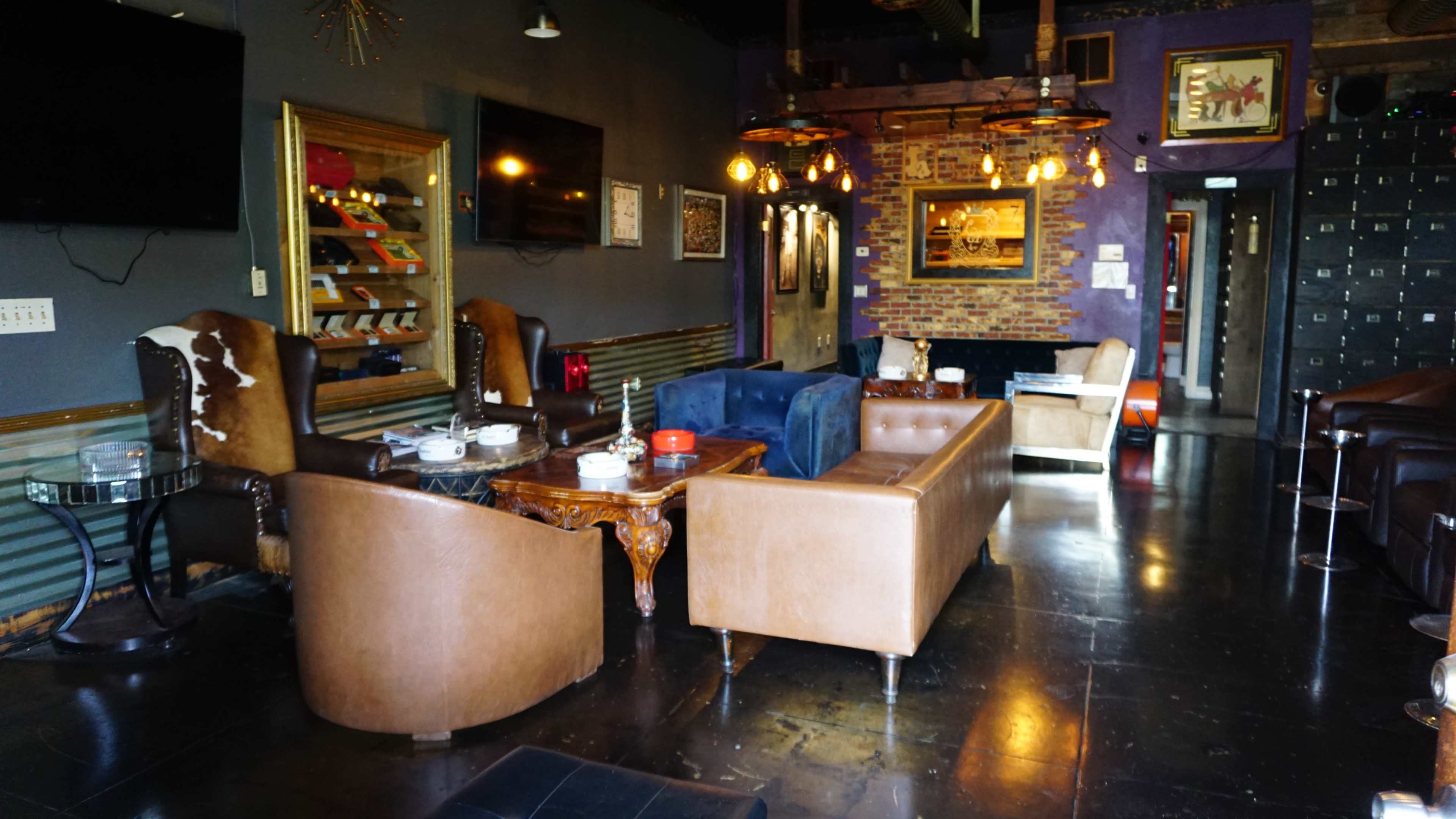 The image shows a stylish lounge area featuring brown leather sofas, a blue velvet couch, wooden tables, and decorative lighting against a dark-themed interior.