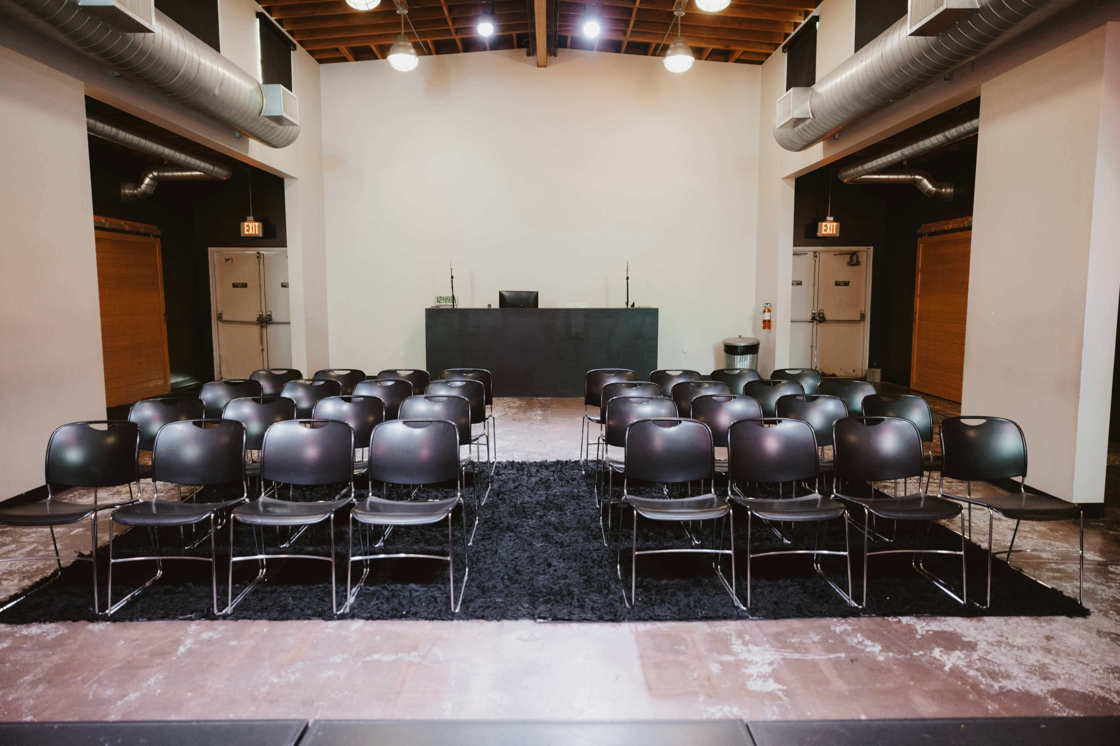 The image shows a spacious room set up with rows of black chairs facing a low stage or platform at the front.