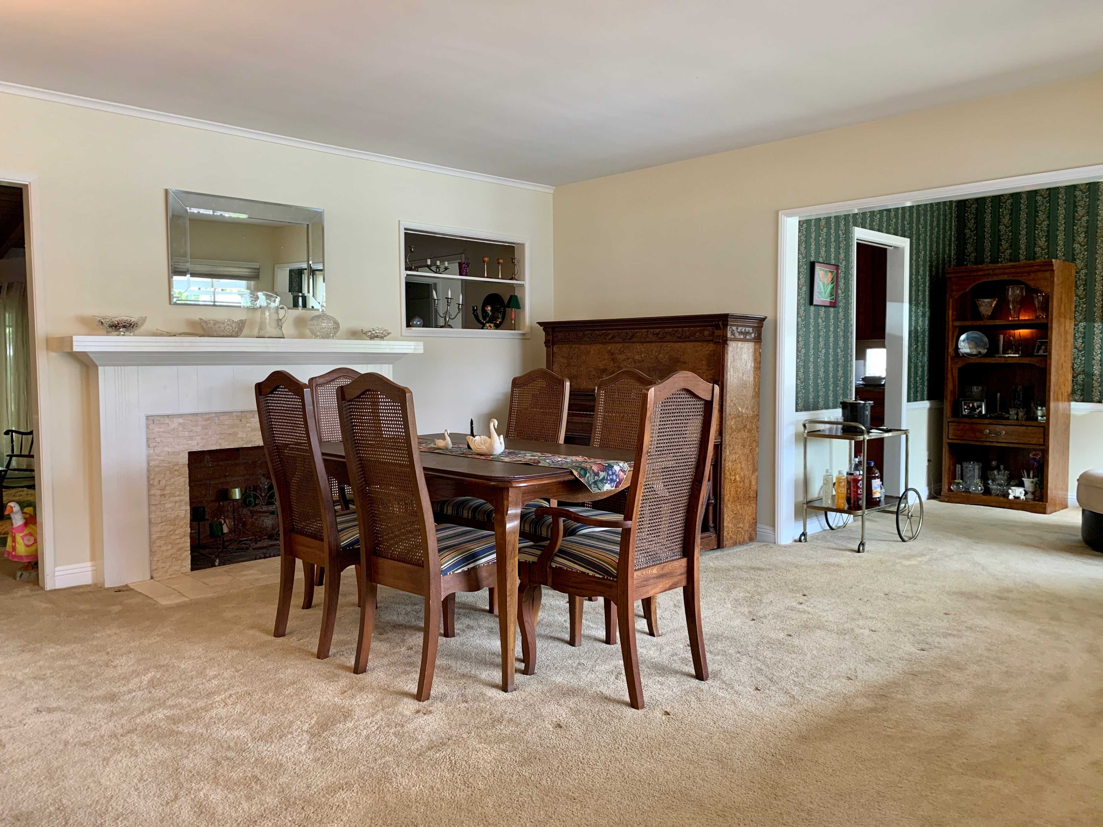 A dining room features a wooden table surrounded by six chairs, with a fireplace and a doorway leading to another room in the background.
