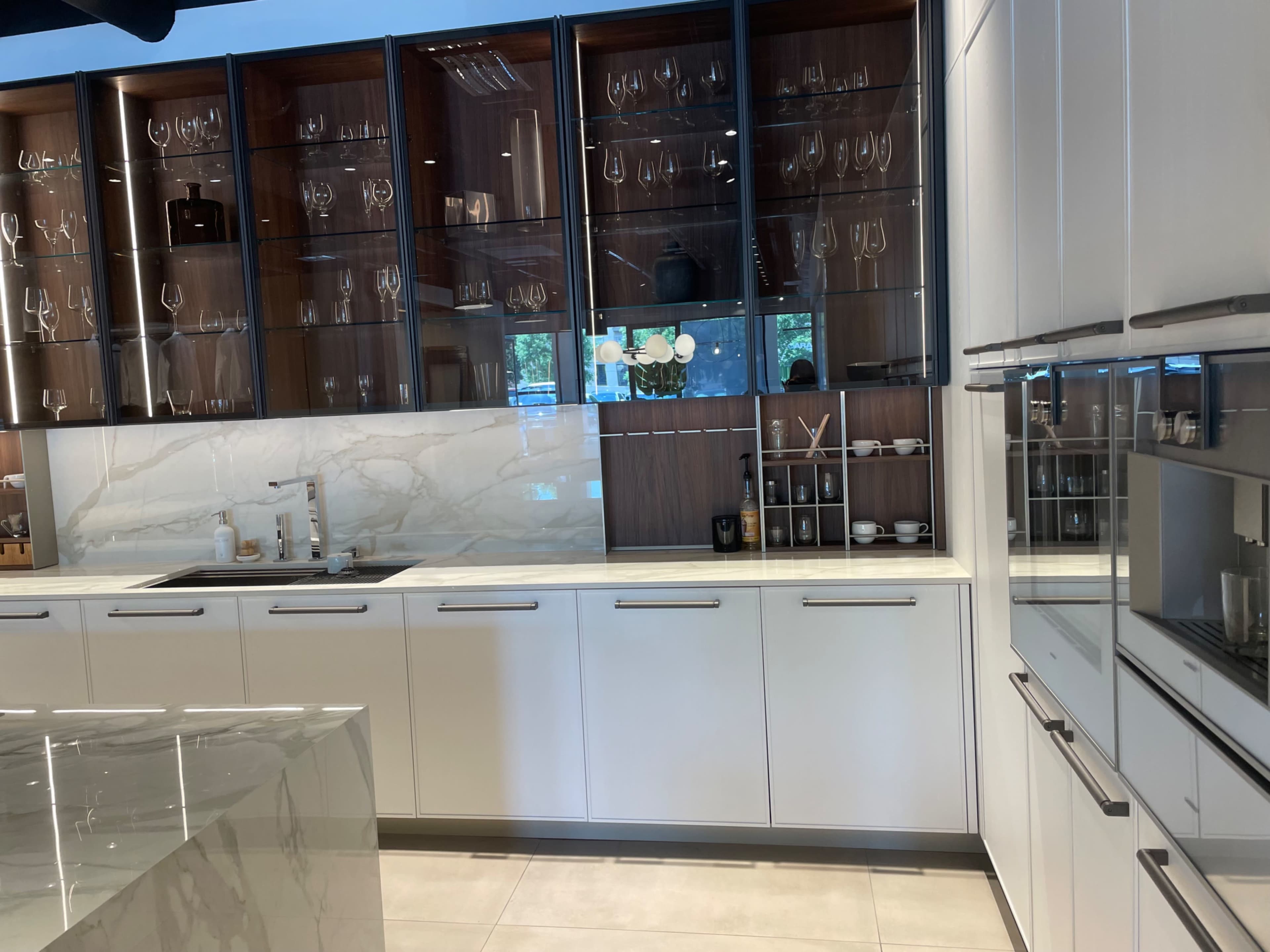 A modern kitchen with sleek white cabinetry, glass-fronted cabinets displaying glassware, and a marble countertop.