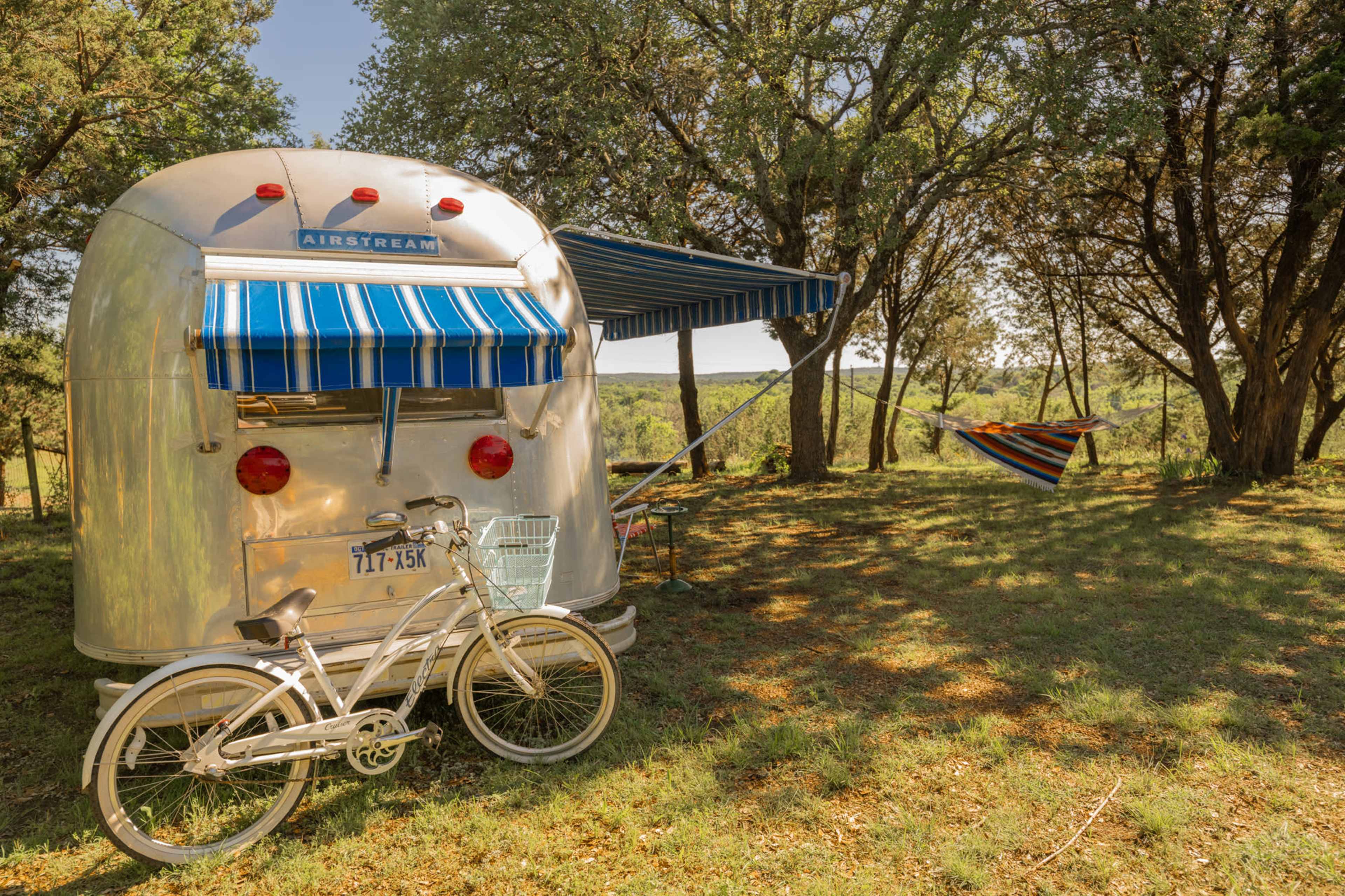 An Airstream trailer with a striped awning is parked under trees, alongside a bicycle and a colorful hammock.