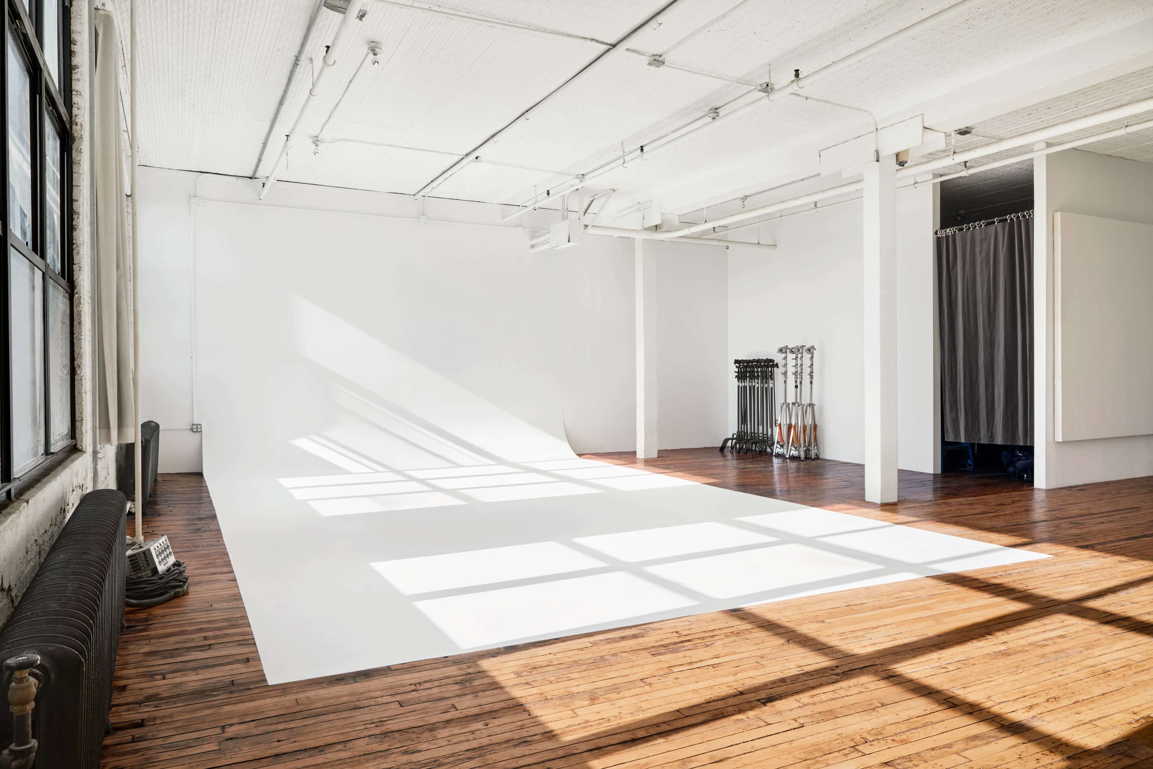 A spacious, empty photography studio with a white backdrop and wooden floors, illuminated by natural light streaming through large windows.