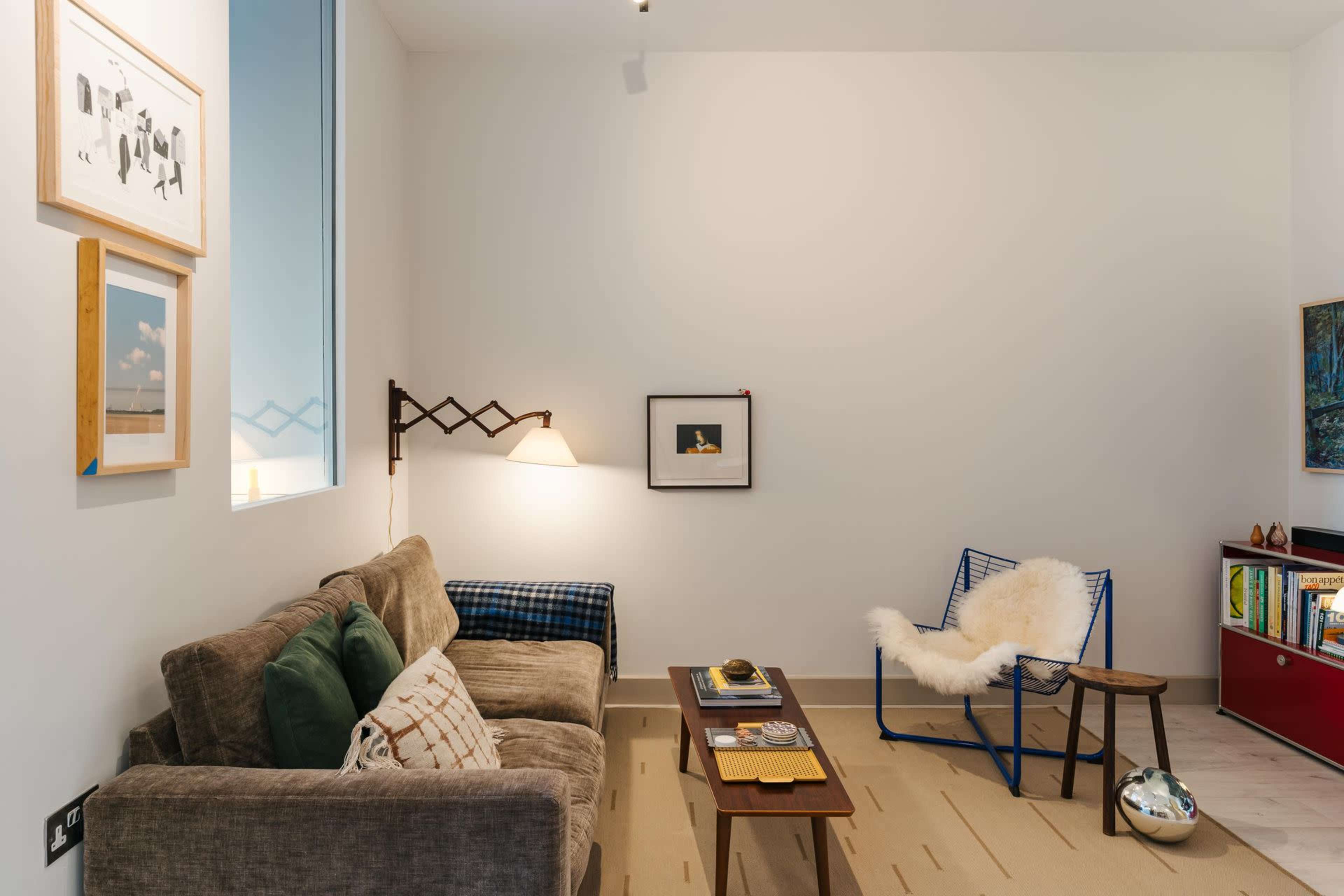 The room features a brown sofa, a minimalist chair, a wooden table with decorative items, and various framed artworks on the walls.