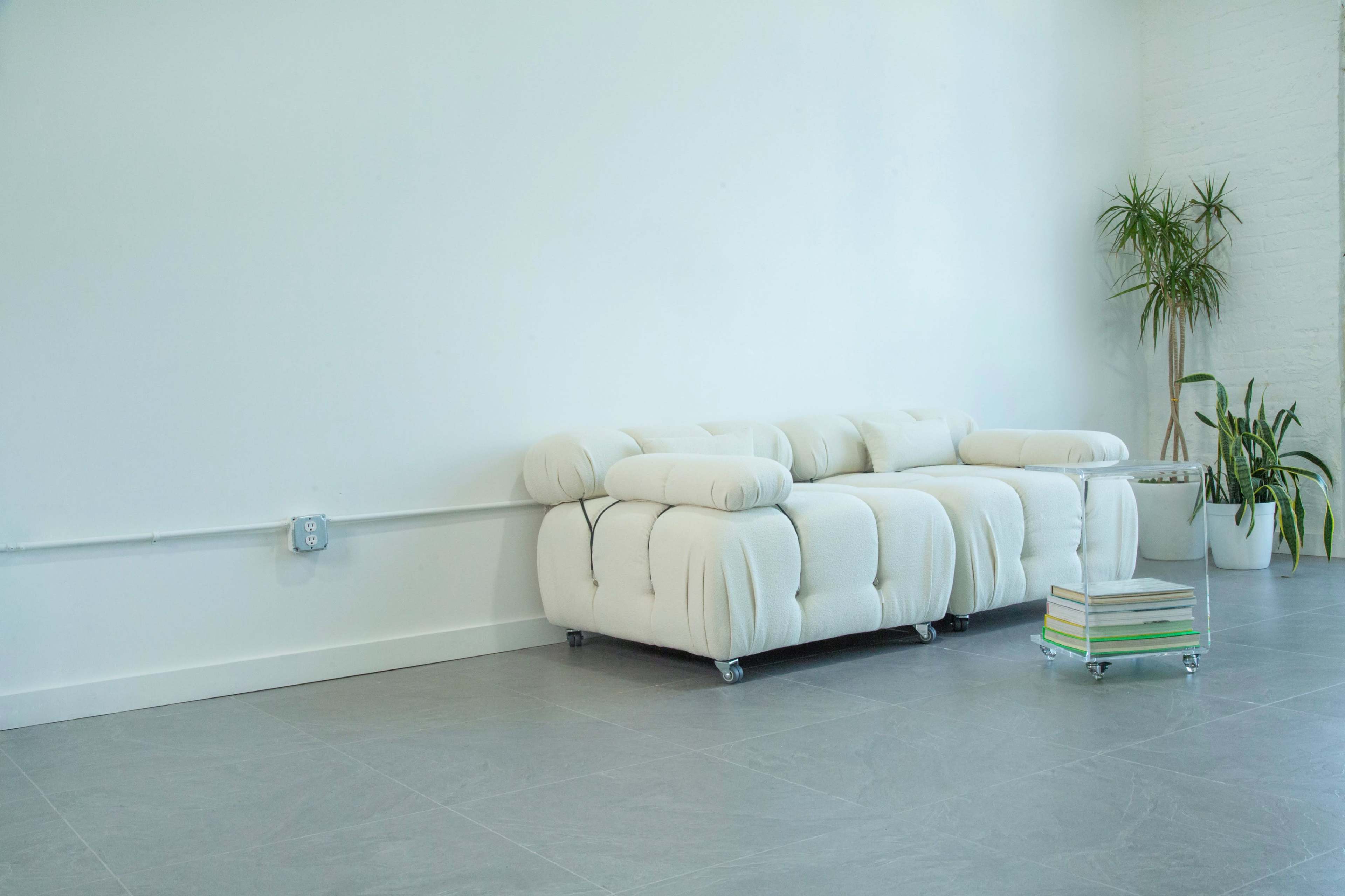 A white, tufted sofa is placed against a blank wall in a spacious room, accompanied by a small table and potted plants.