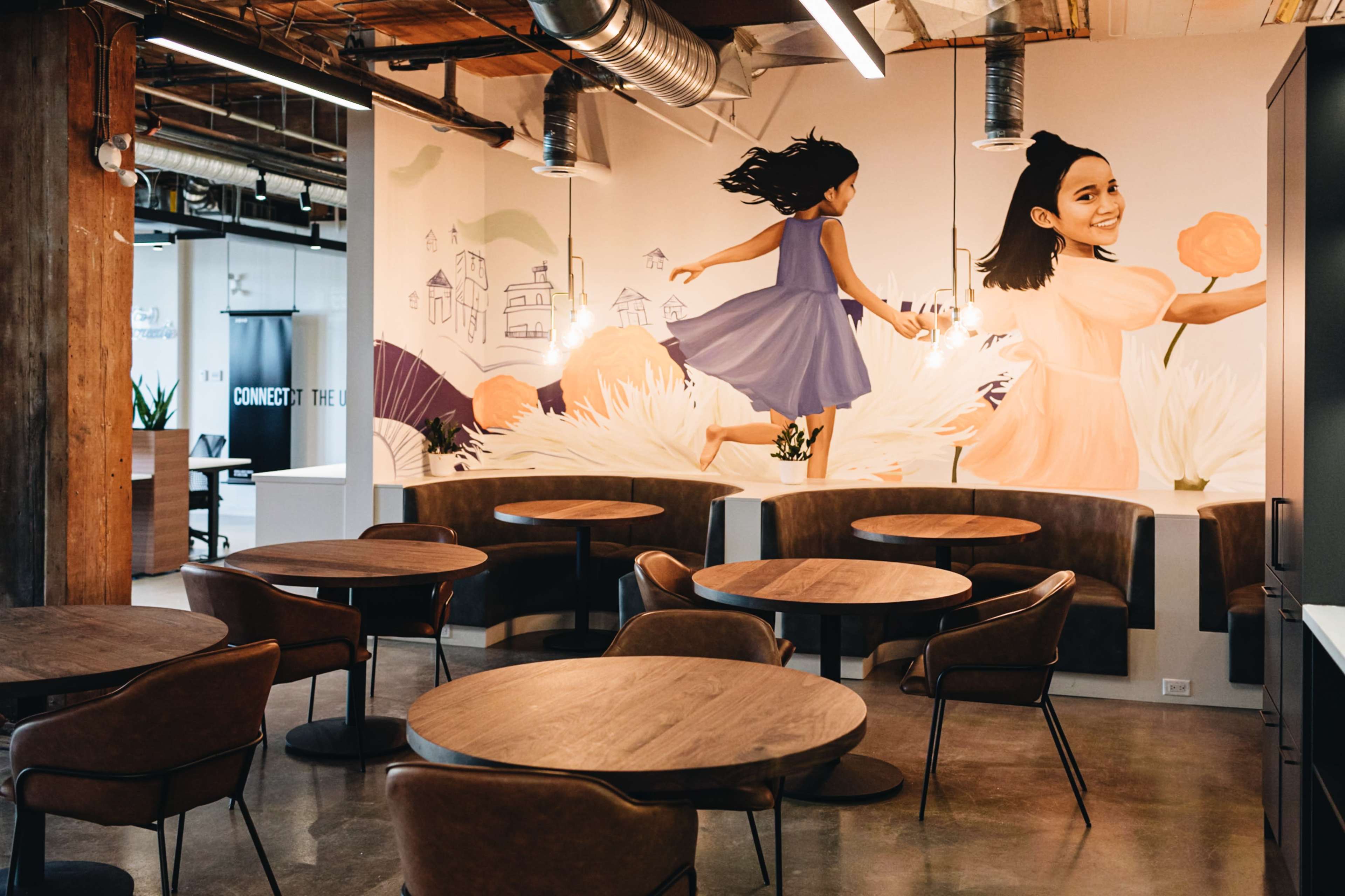 The image shows a modern office space with wooden tables and chairs, featuring a mural of a girl running and another girl holding flowers on the wall.