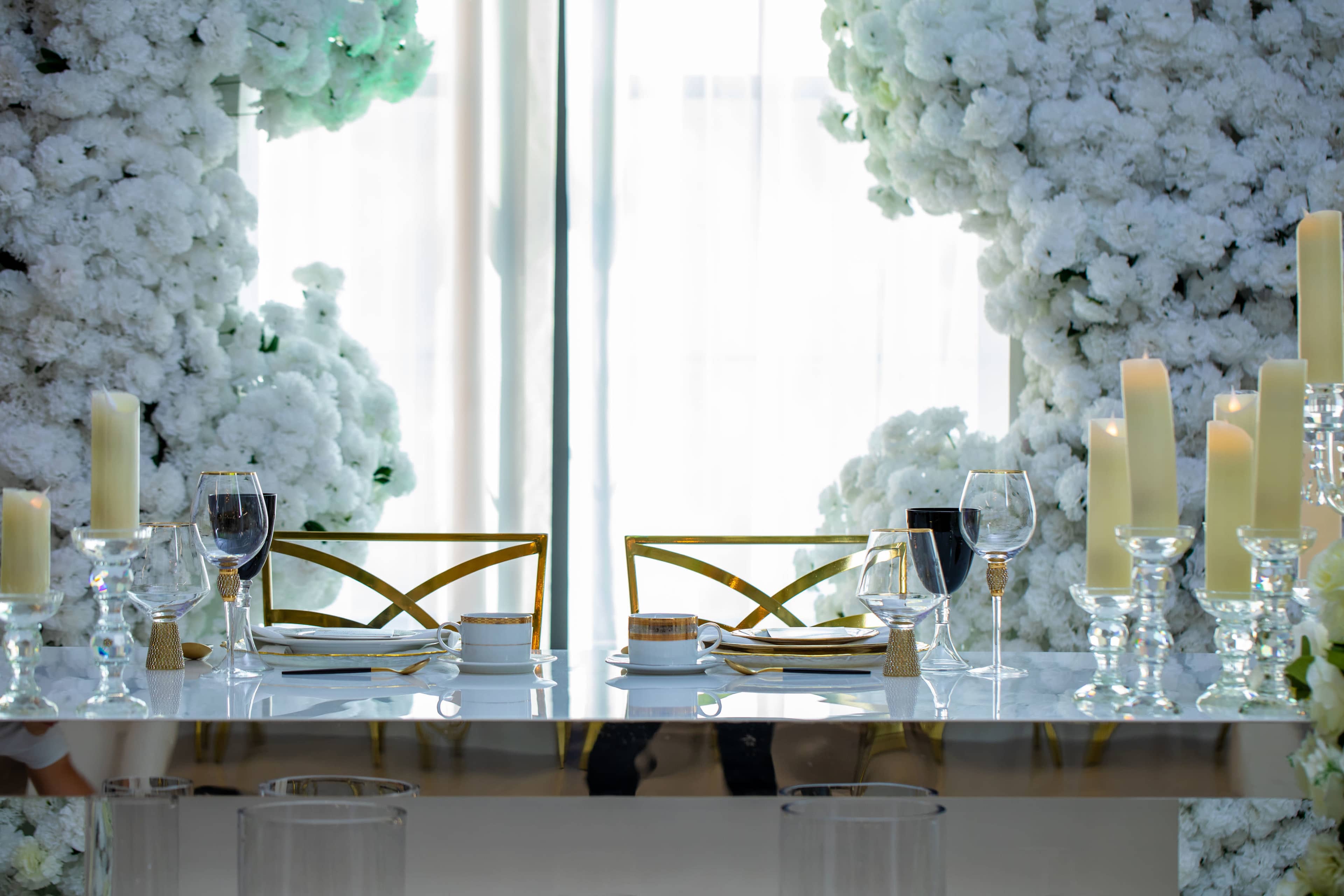 An elegantly set dining table with clear glasses, white plates, and candles, surrounded by tall arrangements of white flowers.