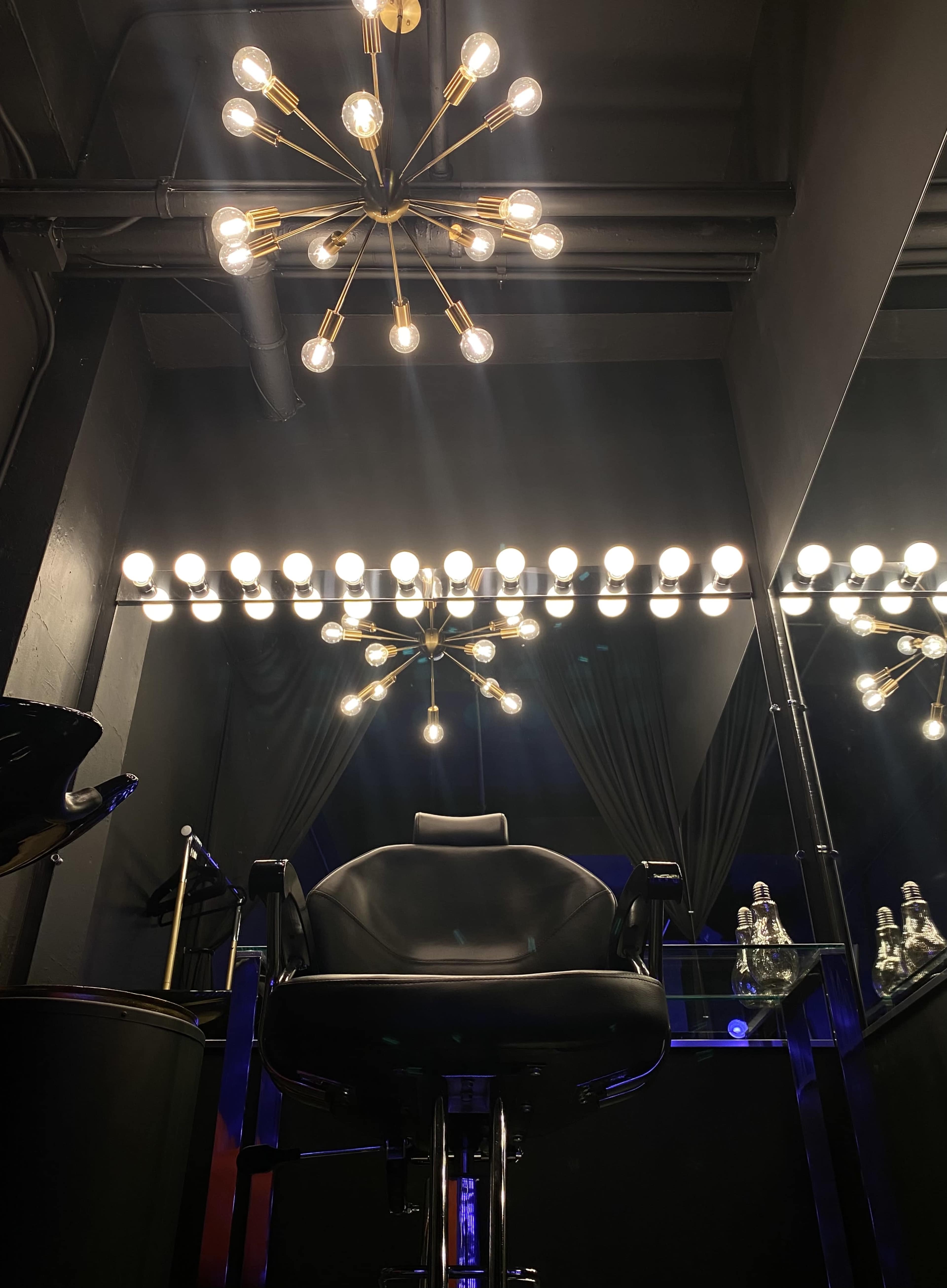 A black barber chair under a stylish light fixture in a dimly lit salon, with a large mirror and illuminated bulbs above.