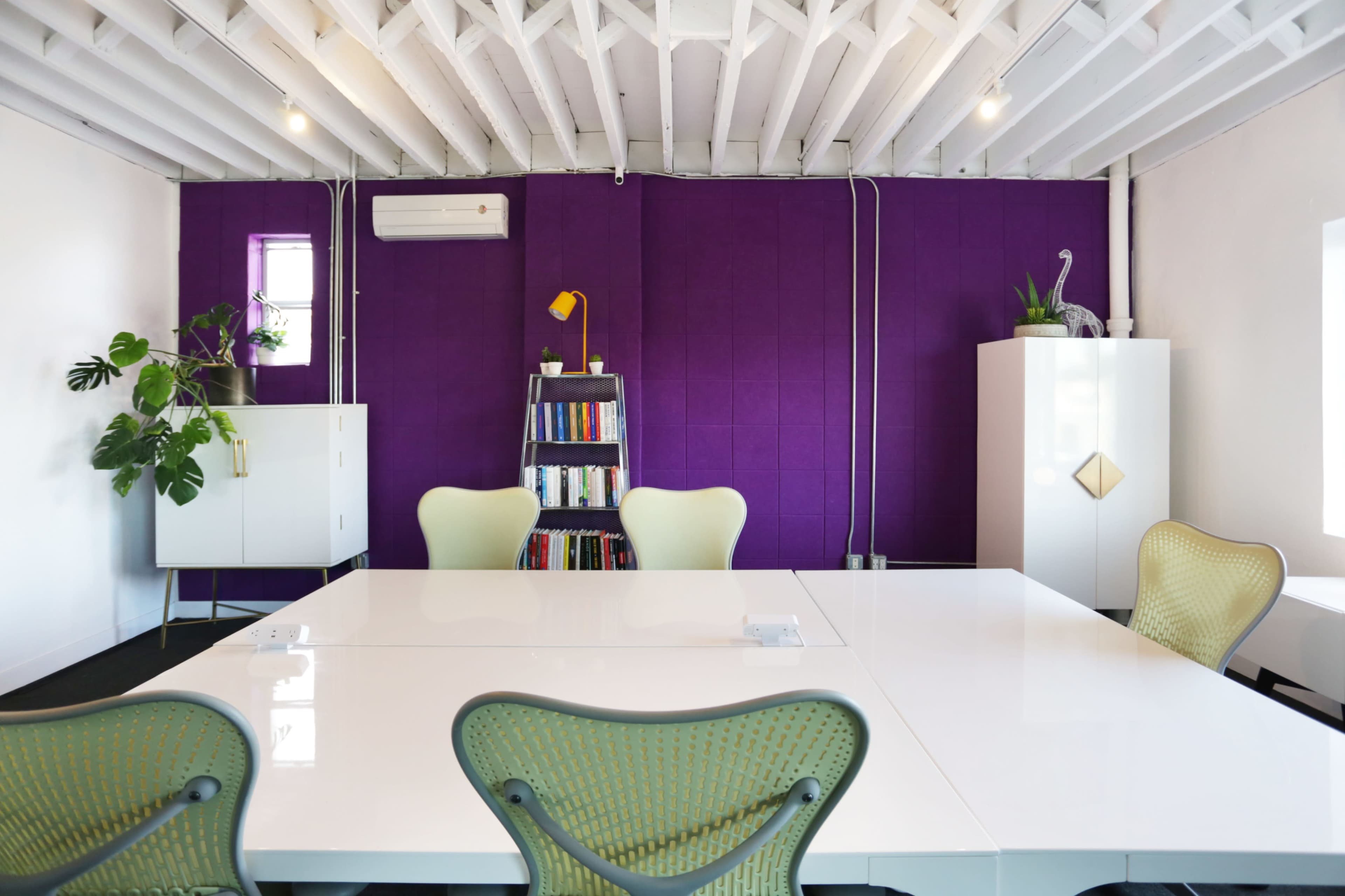 A bright meeting room features a white table, green chairs, a purple accent wall, and shelves with books and plants.