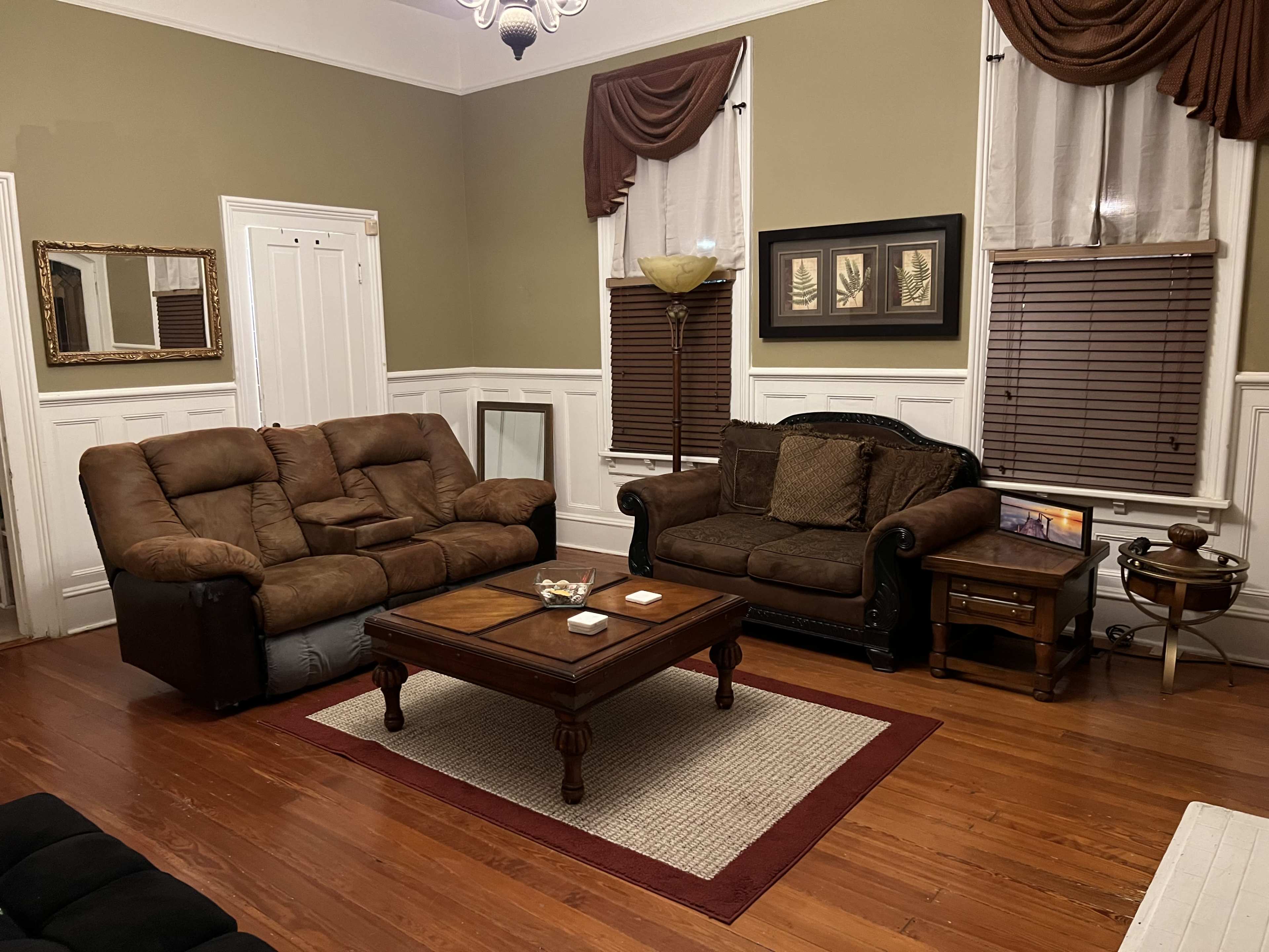 A living room with two sofas, a coffee table, and wooden flooring, framed by brown window treatments and light-colored walls.