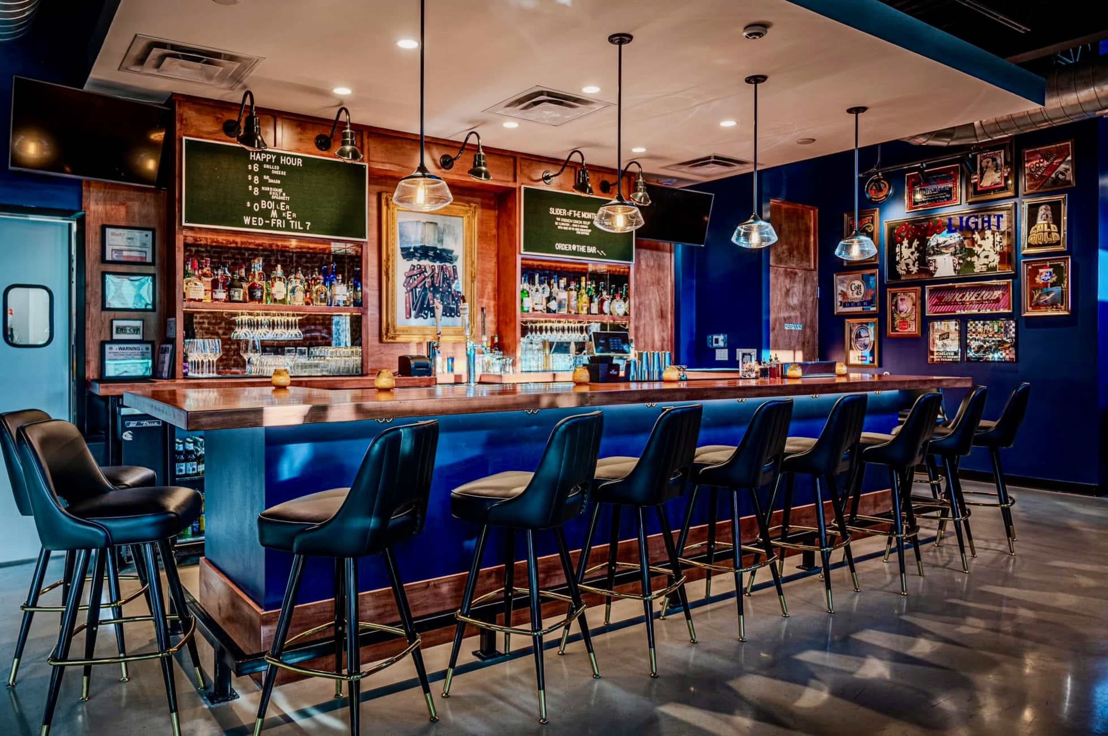 A modern bar features a long wooden counter with high stools, illuminated by pendant lights, surrounded by shelves filled with various bottles and framed artworks on the walls.