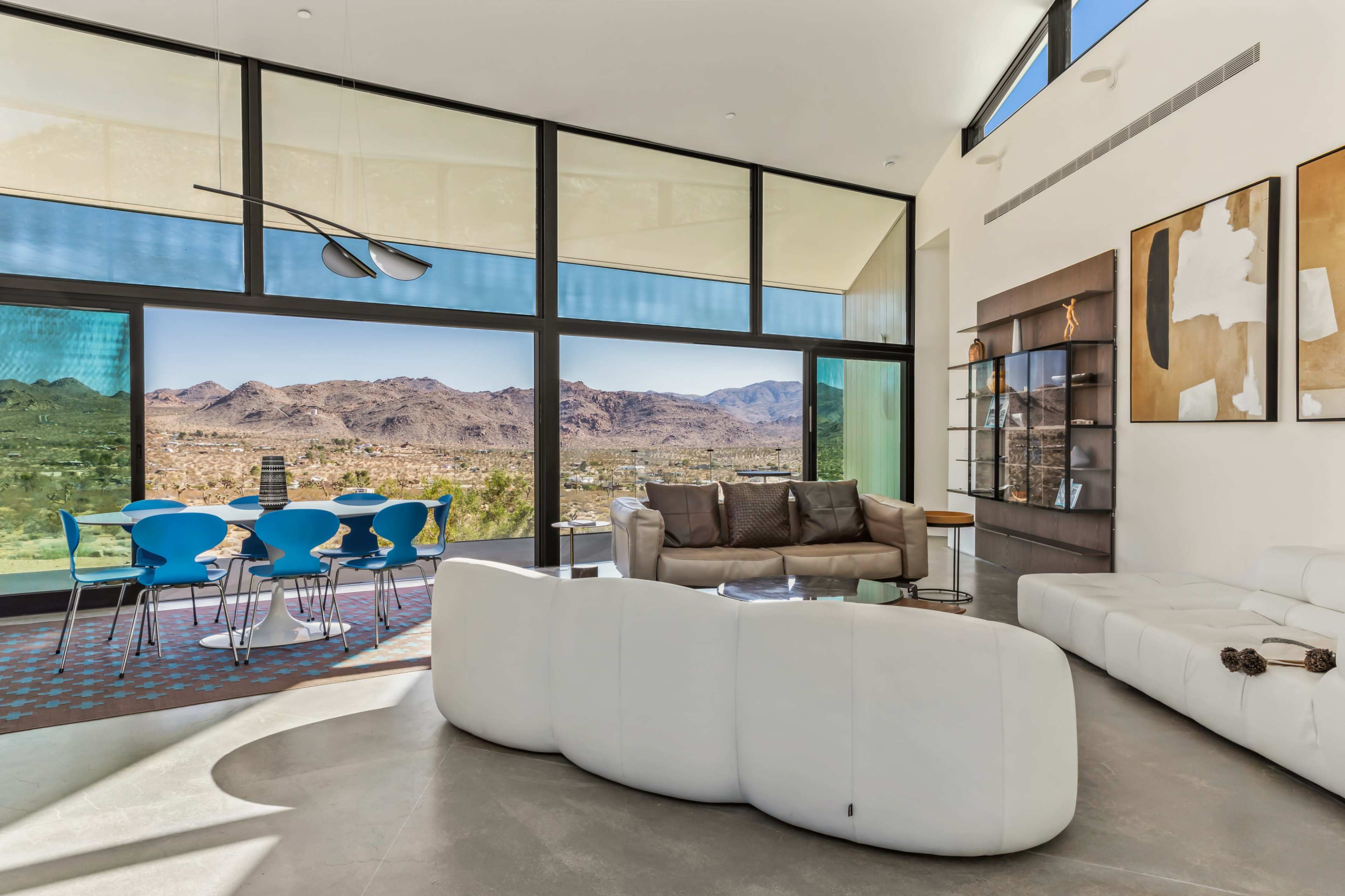 The image shows a modern living space with large windows overlooking a mountainous landscape, featuring contemporary furniture and a dining area with blue chairs.