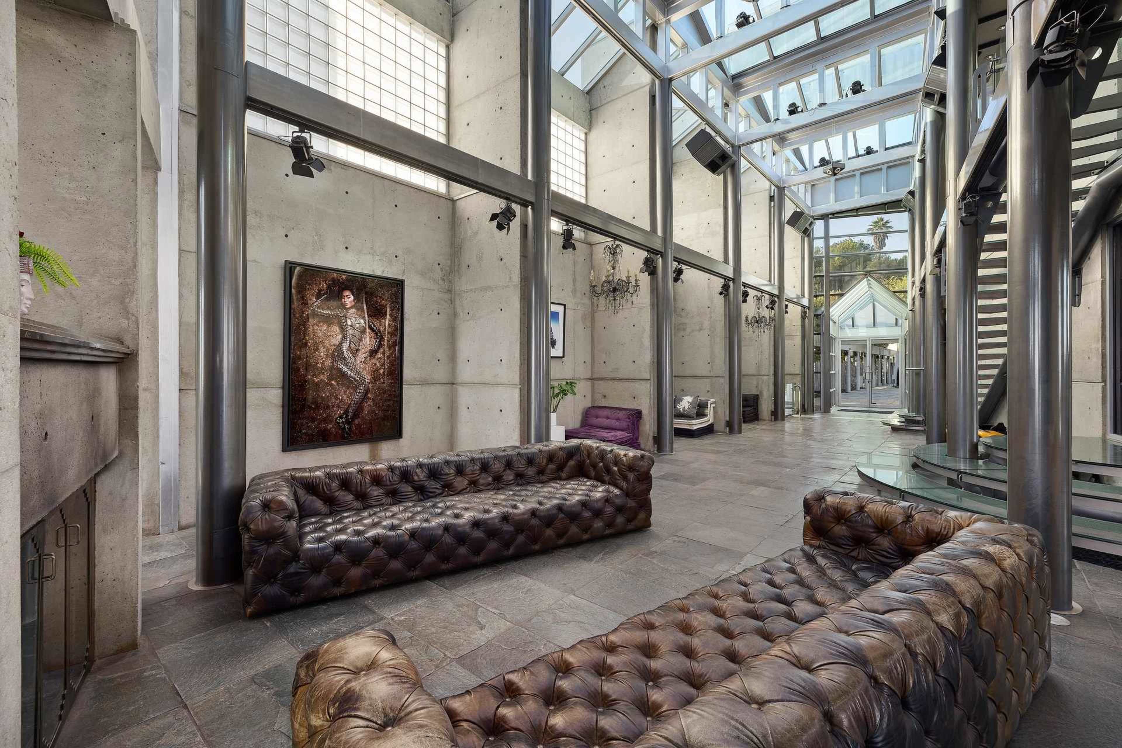 The image features a modern interior space with large glass walls, exposed concrete, and two tufted leather sofas arranged in a bright, open area.