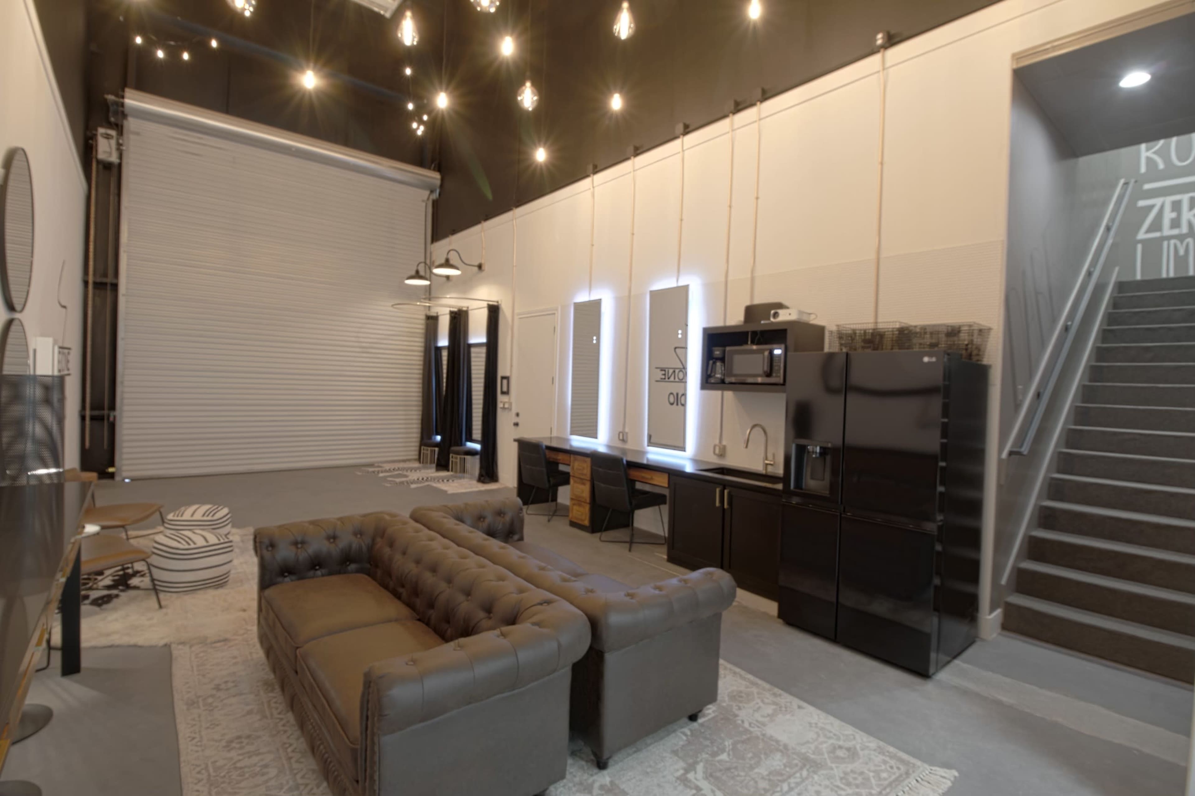 The image shows a spacious interior with modern furniture, including a tufted sofa, a kitchenette with black appliances, and a staircase leading to an upper level.