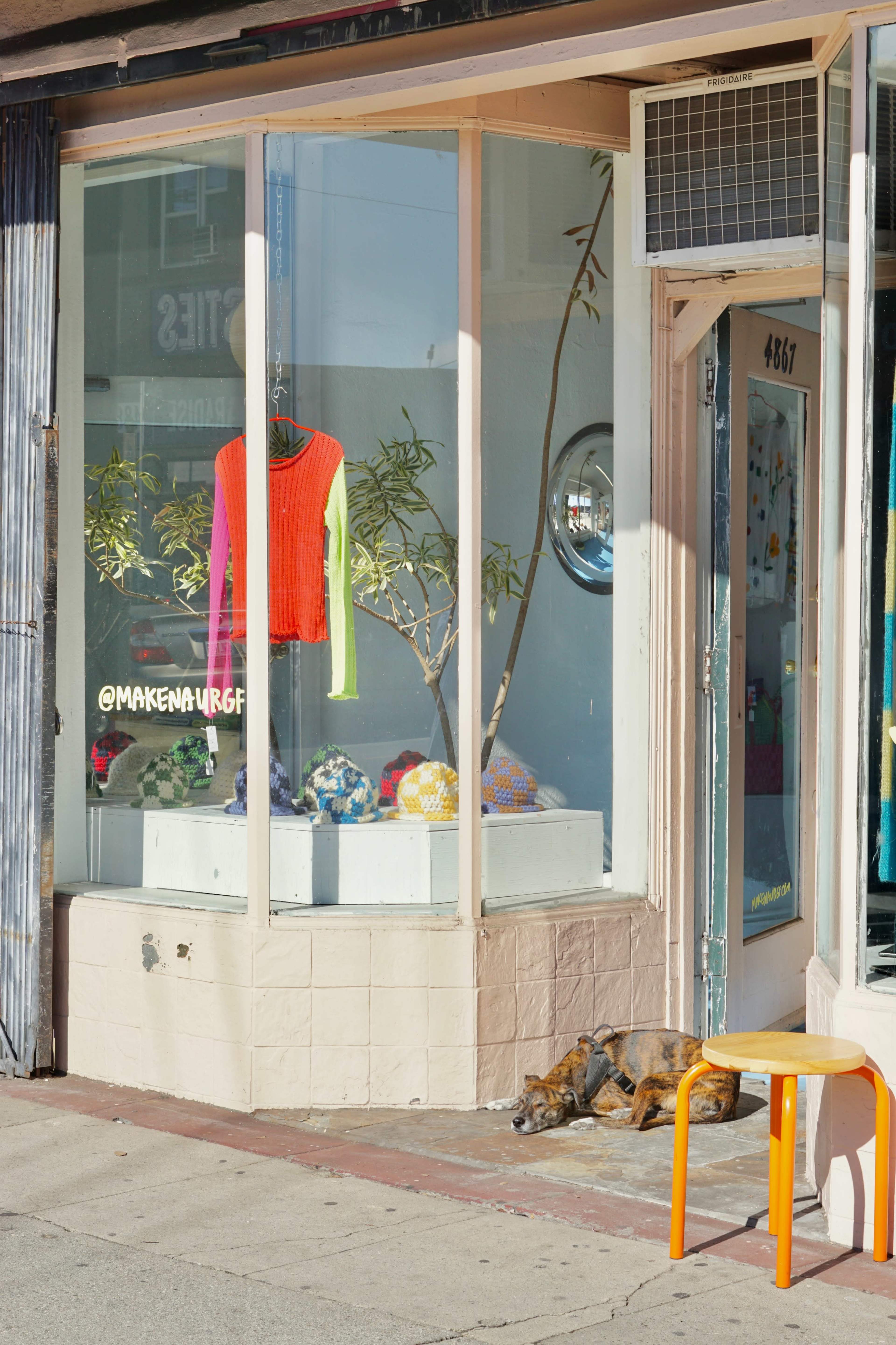 A dog is sleeping on the sidewalk in front of a shop with a bright orange sweater displayed in the window.