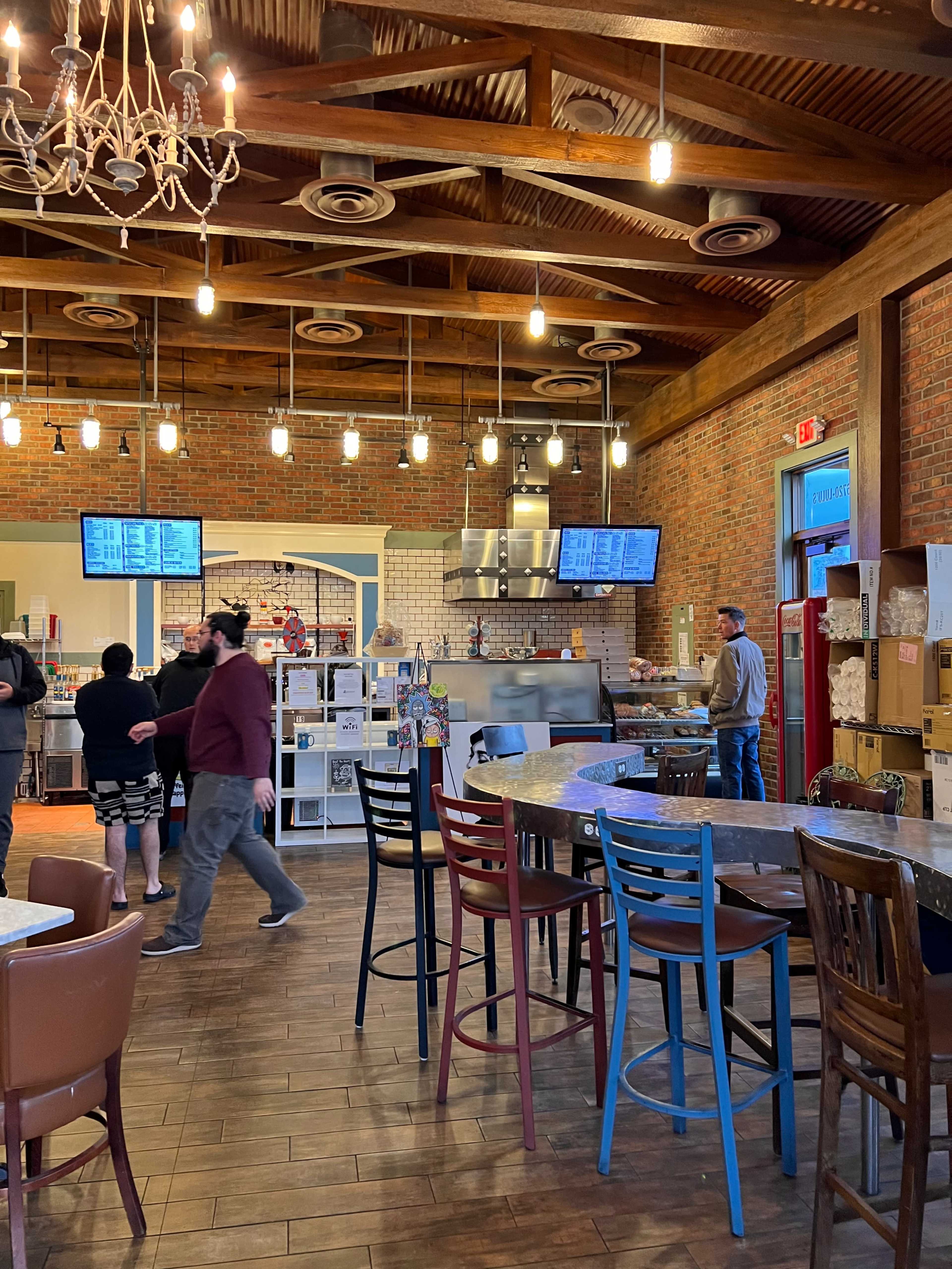 The interior of a modern restaurant features wooden beams, hanging lights, a serving area with menus displayed on screens, and customers seated at various tables and chairs.