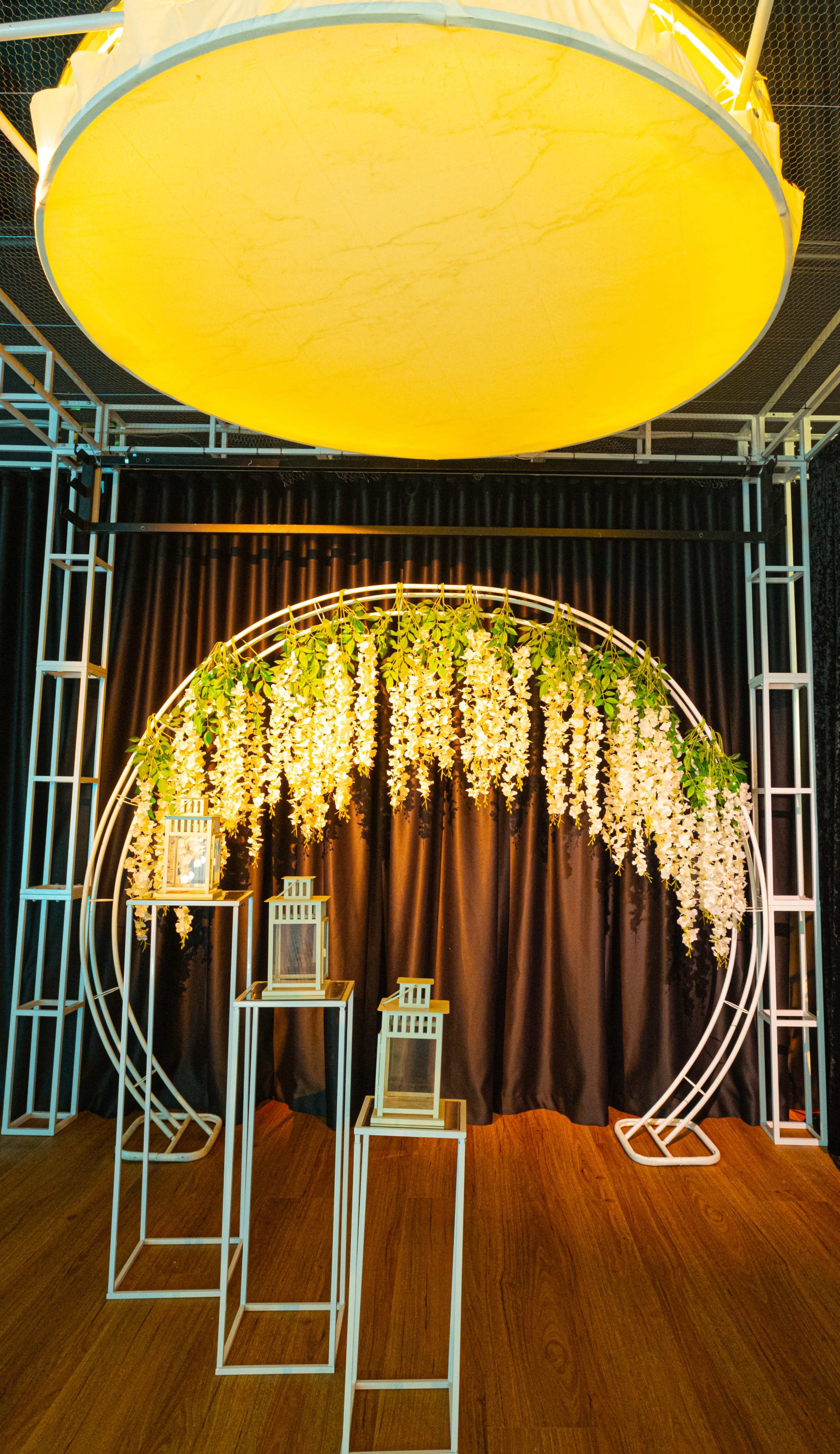 The scene features a circular floral arch draped with hanging flowers, flanked by two pedestal displays, under a yellow-lit circular overhead structure.