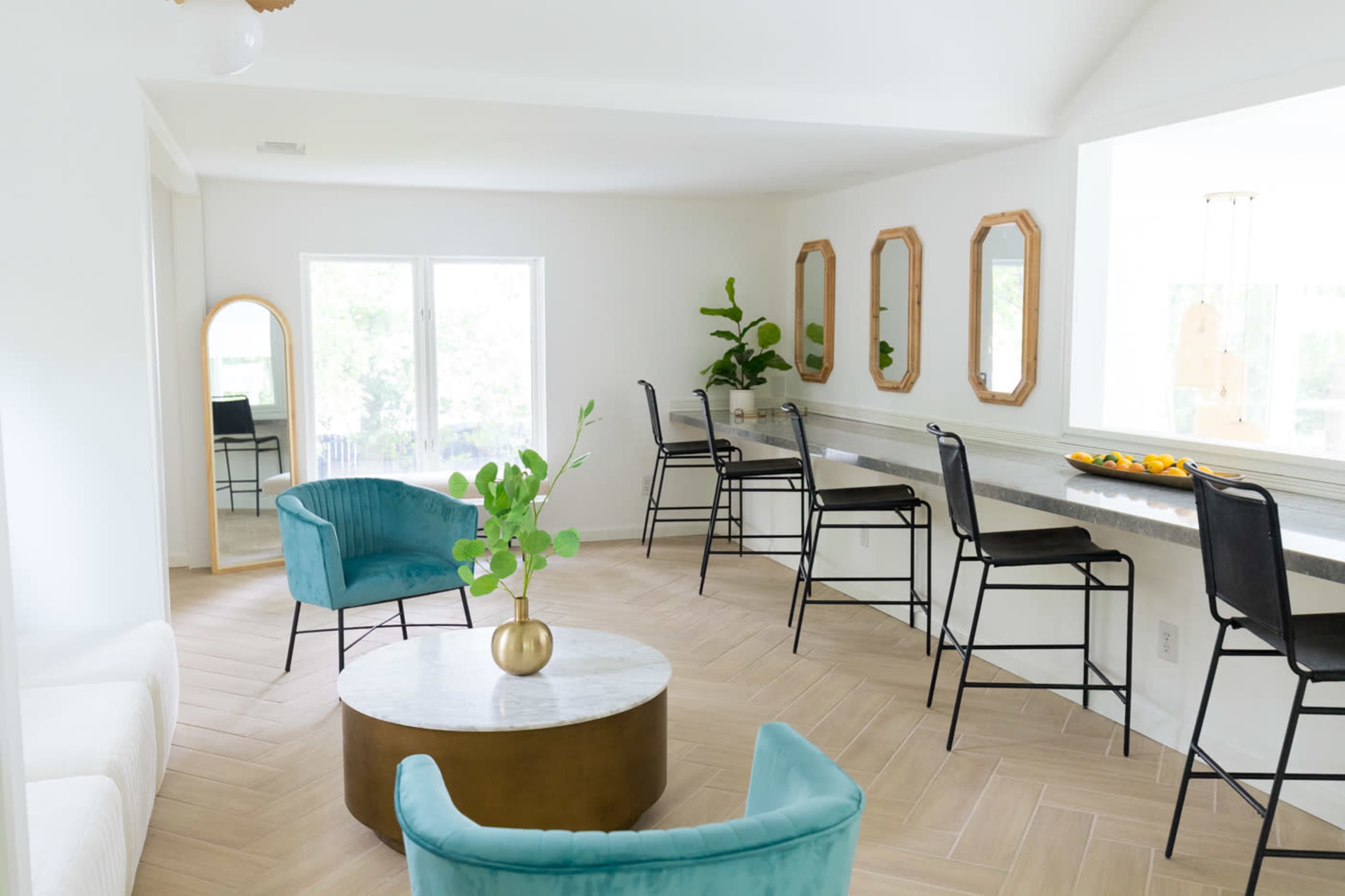 A bright, modern interior features a seating area with two blue chairs, a round coffee table, and a long counter with black chairs and mirrors on the walls.
