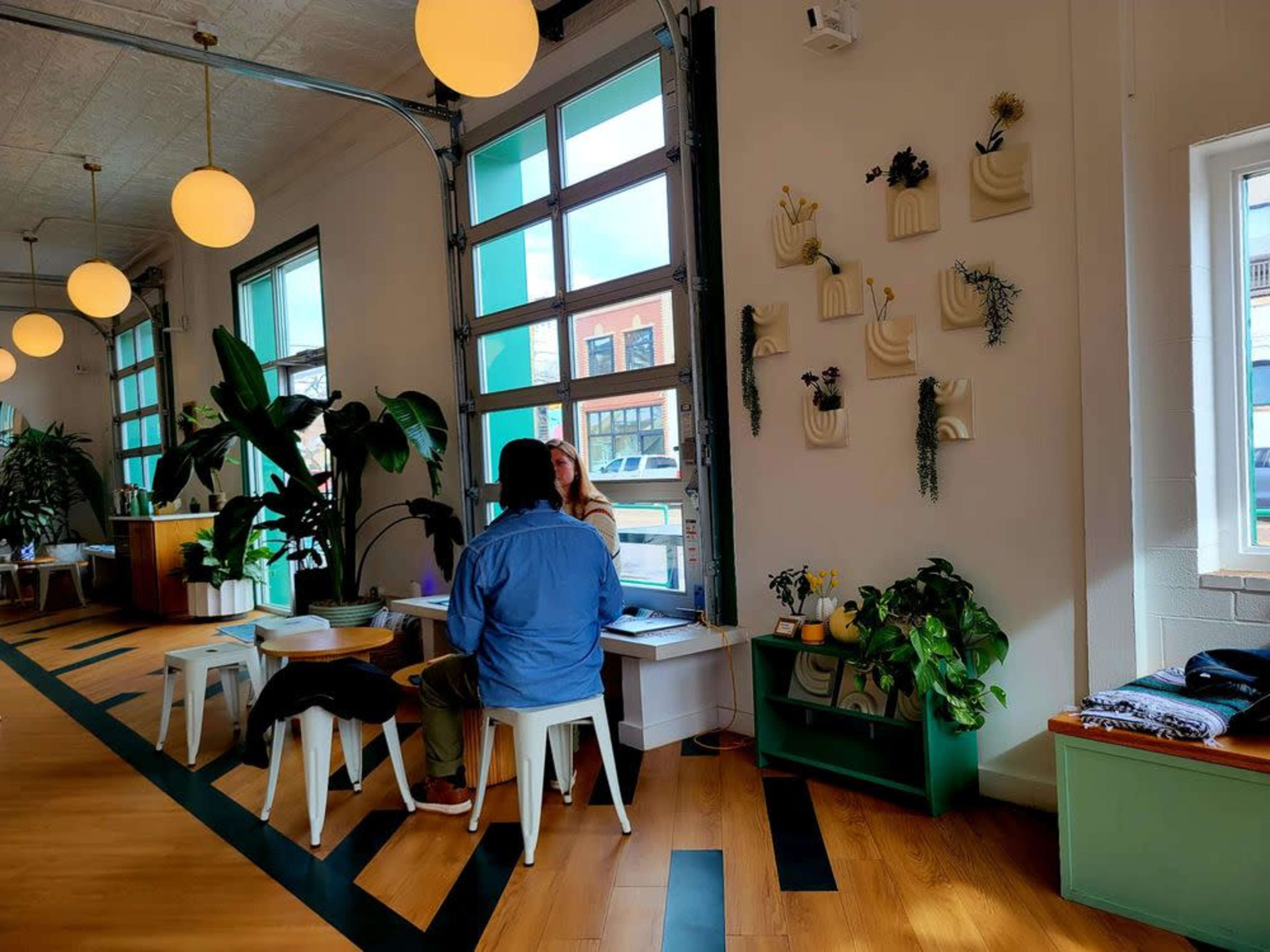 Two people are seated at a table in a brightly lit café, surrounded by plants and minimalist décor, with large windows revealing a view of a street outside.