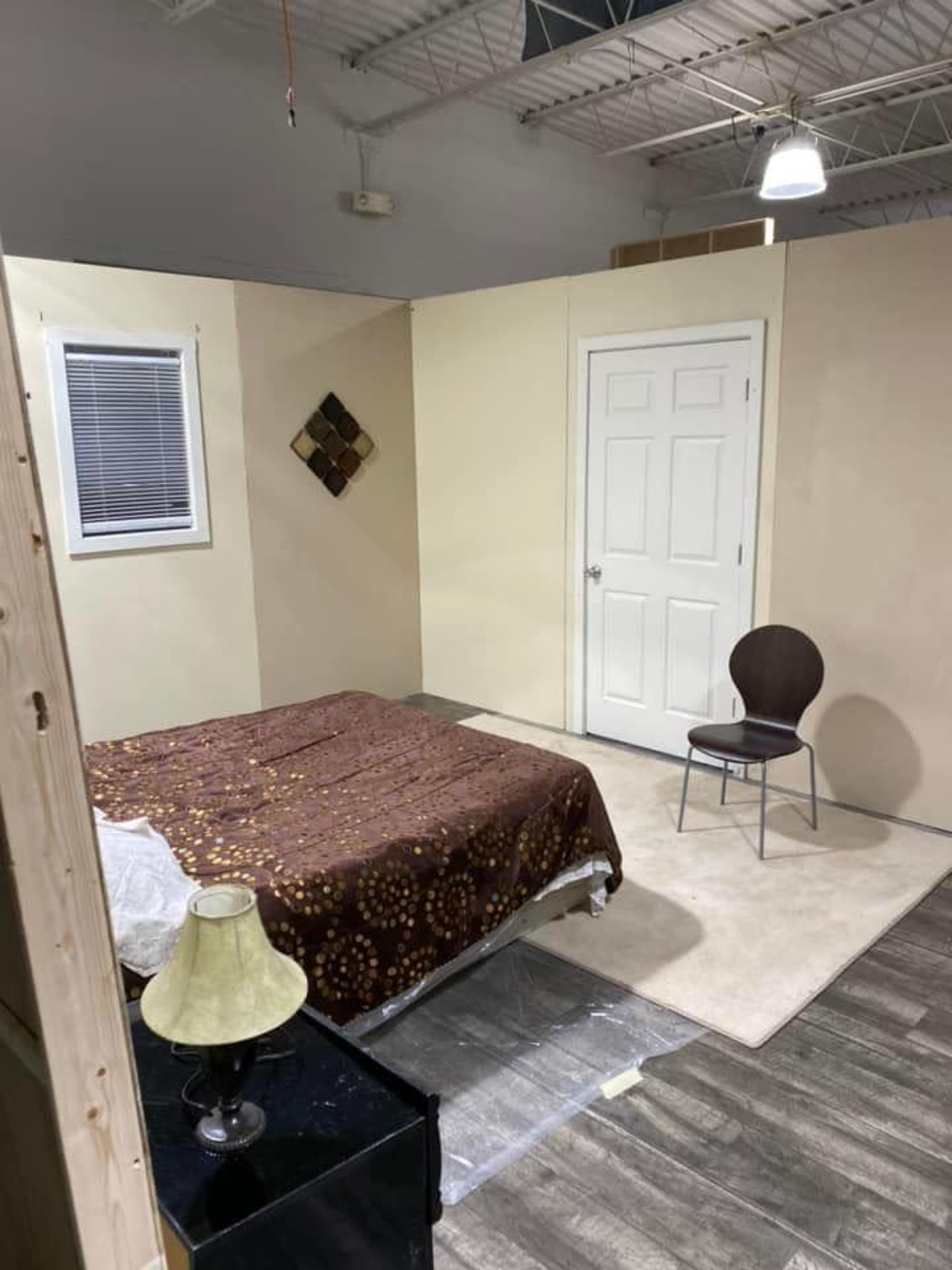The image shows a simple bedroom setup with a bed covered in a brown patterned blanket, a chair, and a lamp beside a nightstand in a lightly colored room.