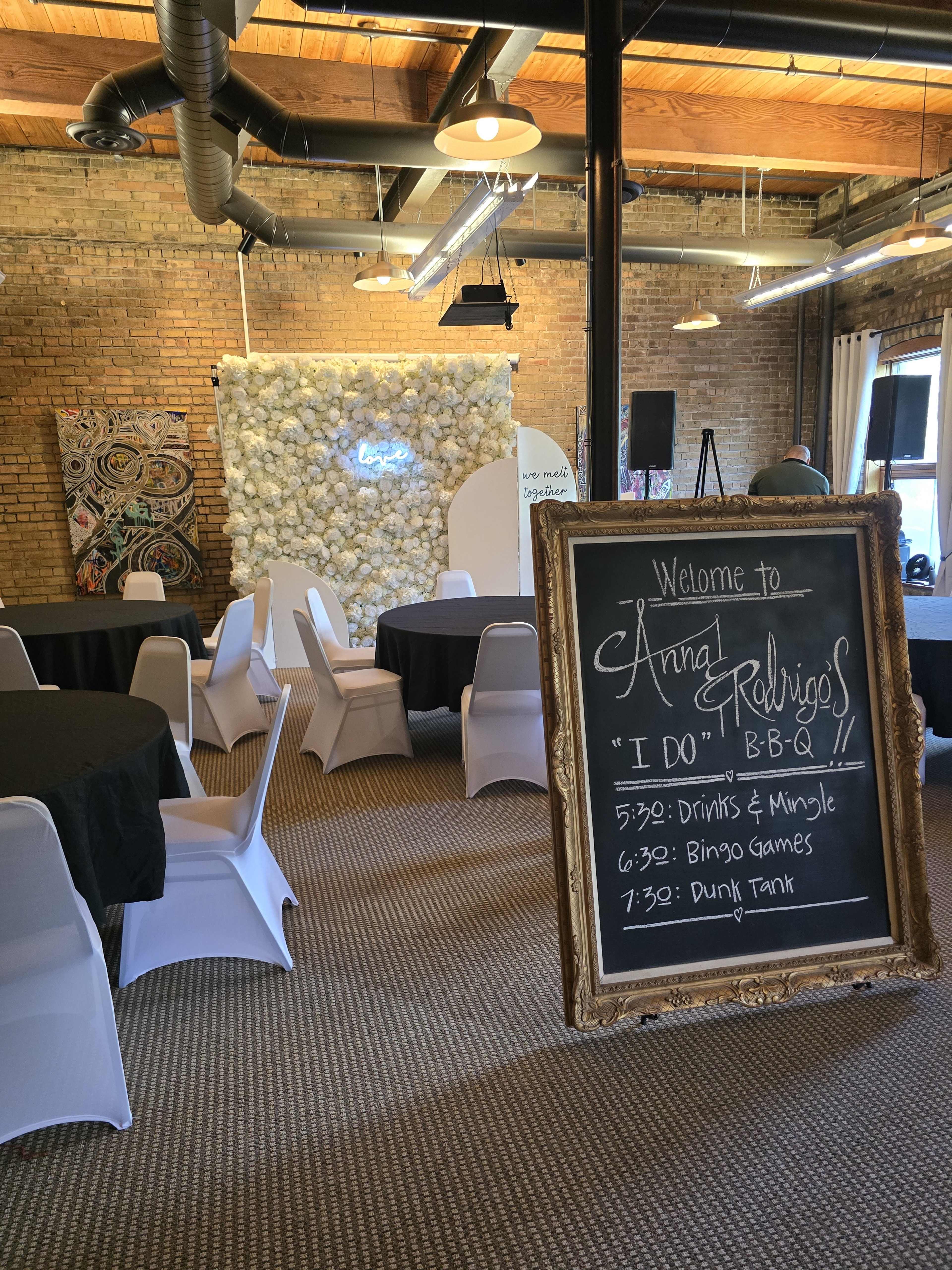 An event space with round tables covered in black cloths, white chairs, and a decorative wall made of flowers, alongside a welcome sign detailing the schedule for a BBQ event.